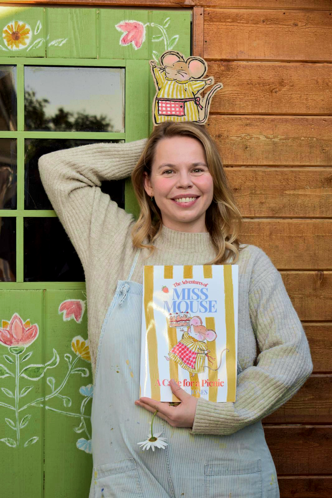 A woman with shoulder-length blonde hair wearing a beige sweater and light-colored apron, standing in front of a wooden wall with green painted wood panels and floral designs. She is smiling and holding a book titled 'The Adventures of Miss Mouse: A Cup for a Picnic' with a picture of a mouse in a yellow-striped dress holding a cake, and she has a daisy flower hanging from her hand.