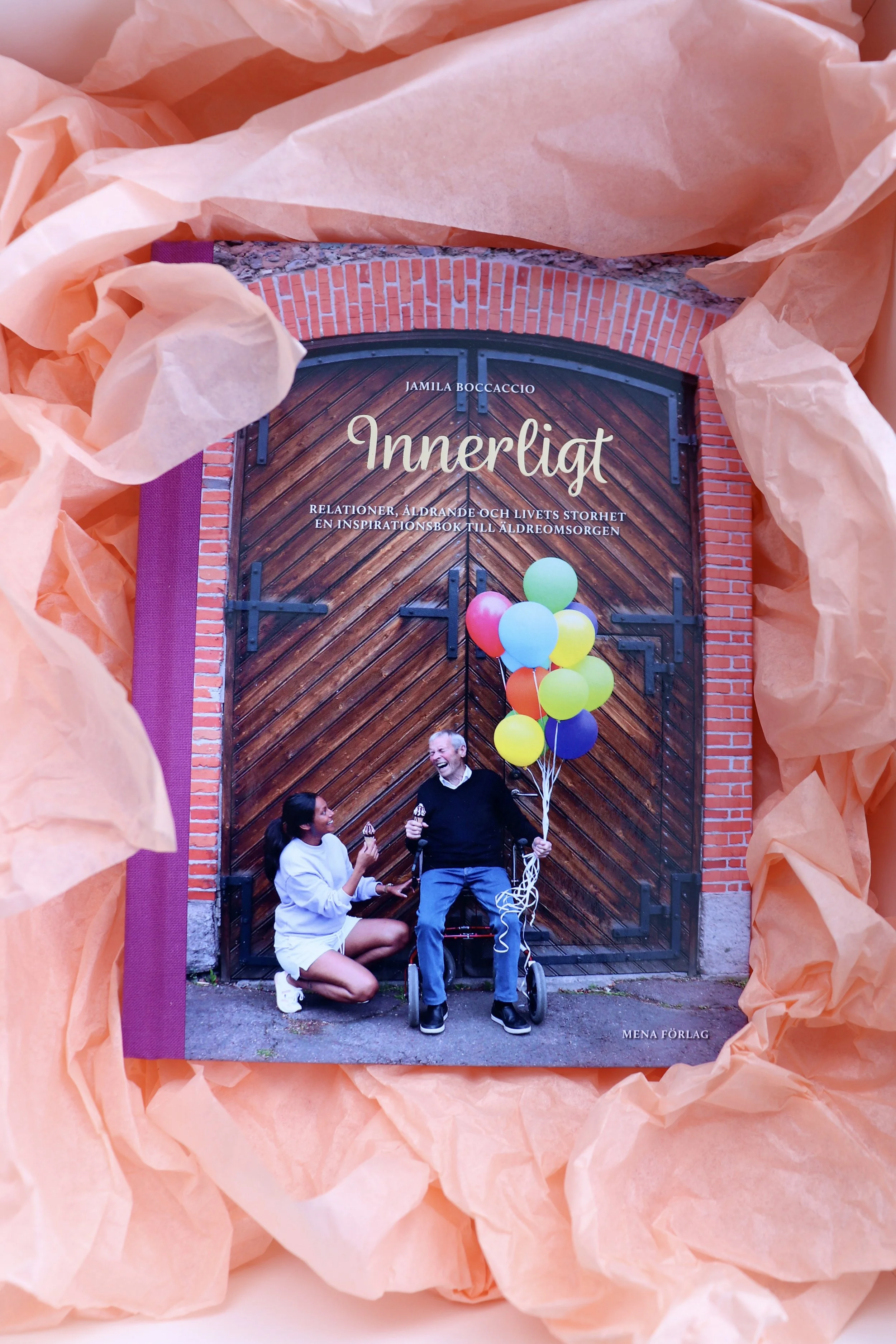 En man och en ung kvinna står framför en stor, träport. Mannen håller en bunt färgglada ballonger och en rullstol. De verkar prata glatt med varandra. Boken på bilden heter 'Innerligt' av Jamila Boccaccio.