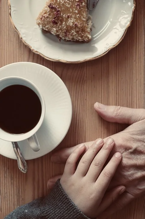 En kopp svart kaffe, ett stycke tårta med kokosflarn, och en vuxen hand som håller ett barns hand på ett träbord.