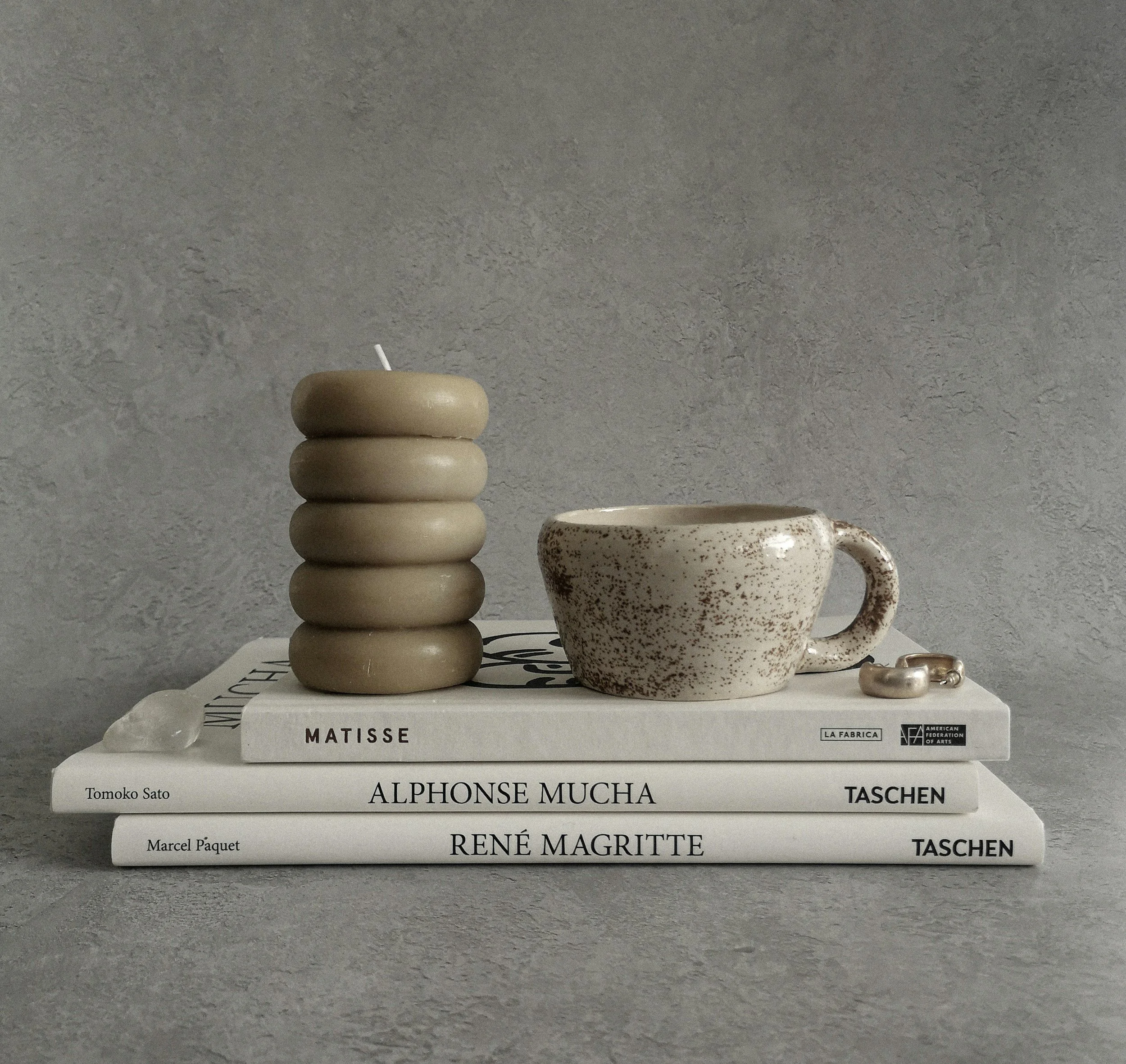 A stack of six beige ceramic bowls on top of several large art books with black and white text on the spines. The books are titled 'Matisse', 'Alphonse Mucha', and 'René Magritte', among others. A ceramic mug with a speckled brown and white pattern and a handle sits to the right of the stack. The background is a textured gray wall.