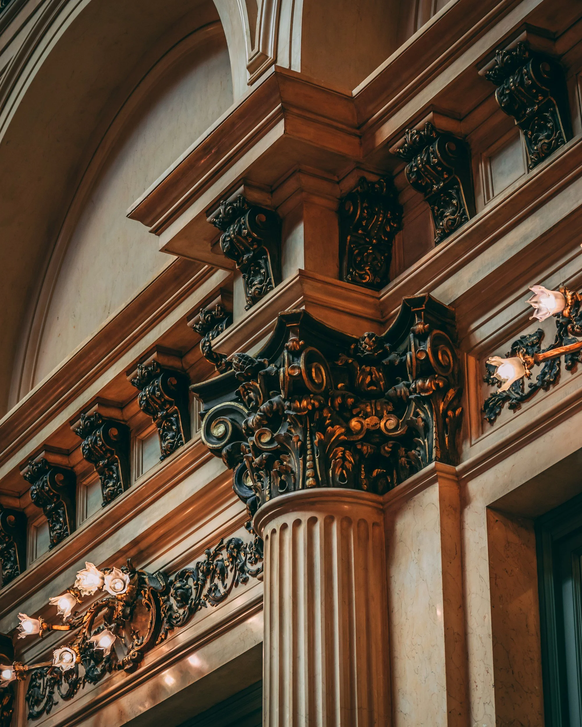 A close-up view of an elaborate wooden clock face with intricate carvings and ornate decorative elements, set against a marble wall and fluted column.