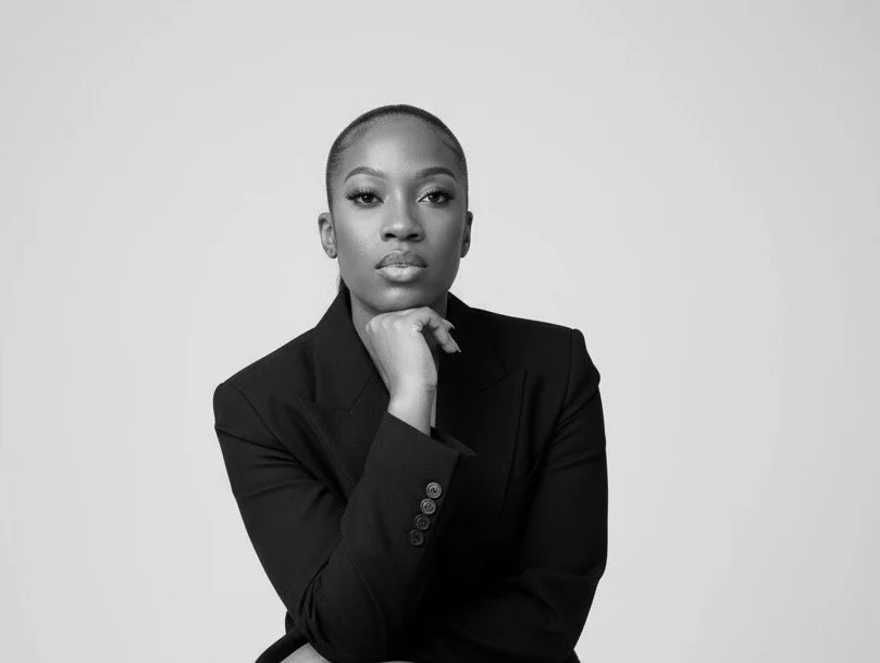 Portrait of a confident woman with a shaved head, wearing a black blazer, resting her chin on her hand, against a plain background.
