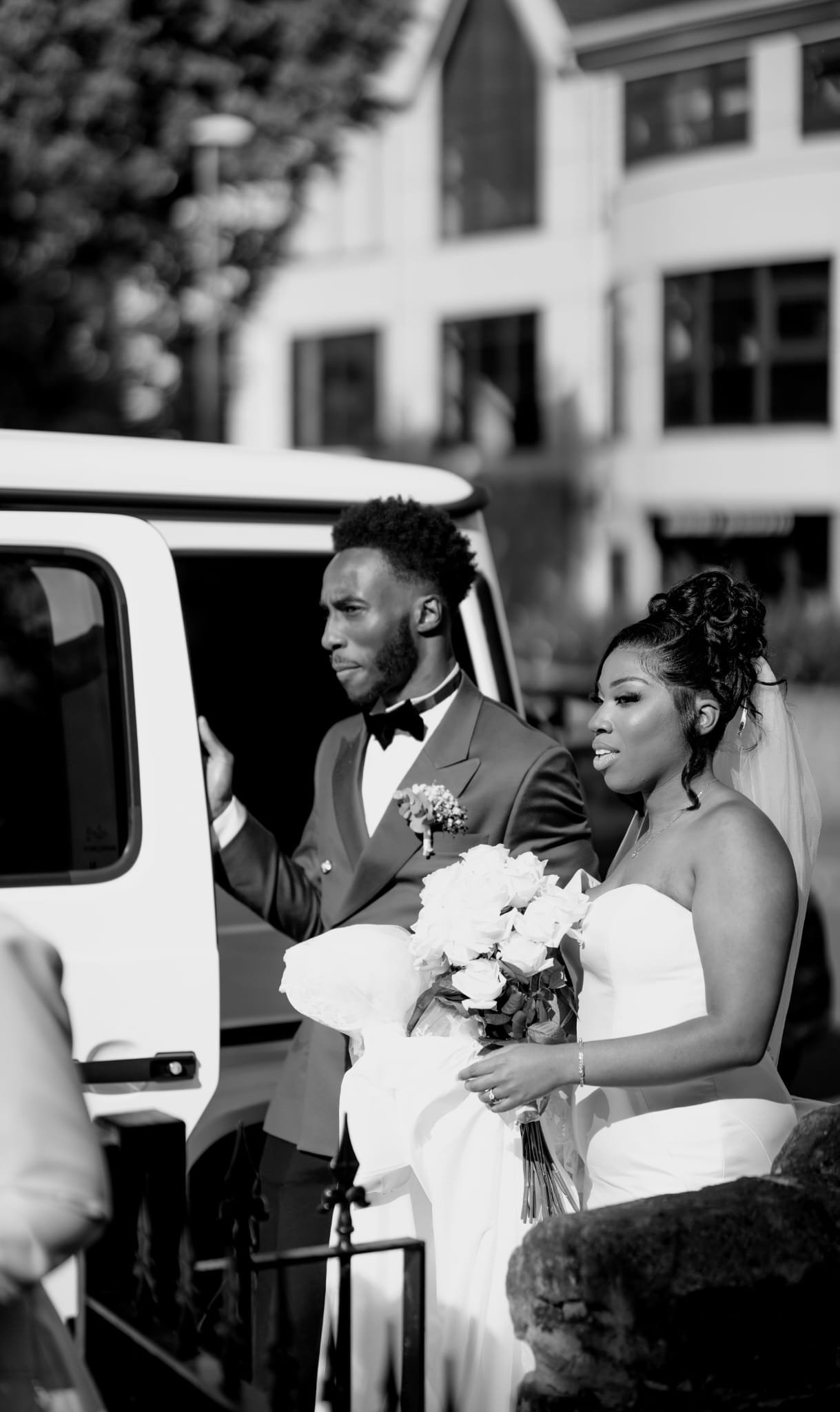 A bride and groom standing outside near a white vehicle. The groom is in a tuxedo and the bride in a strapless wedding dress holding a bouquet of flowers. They appear to be part of a wedding ceremony or celebration.