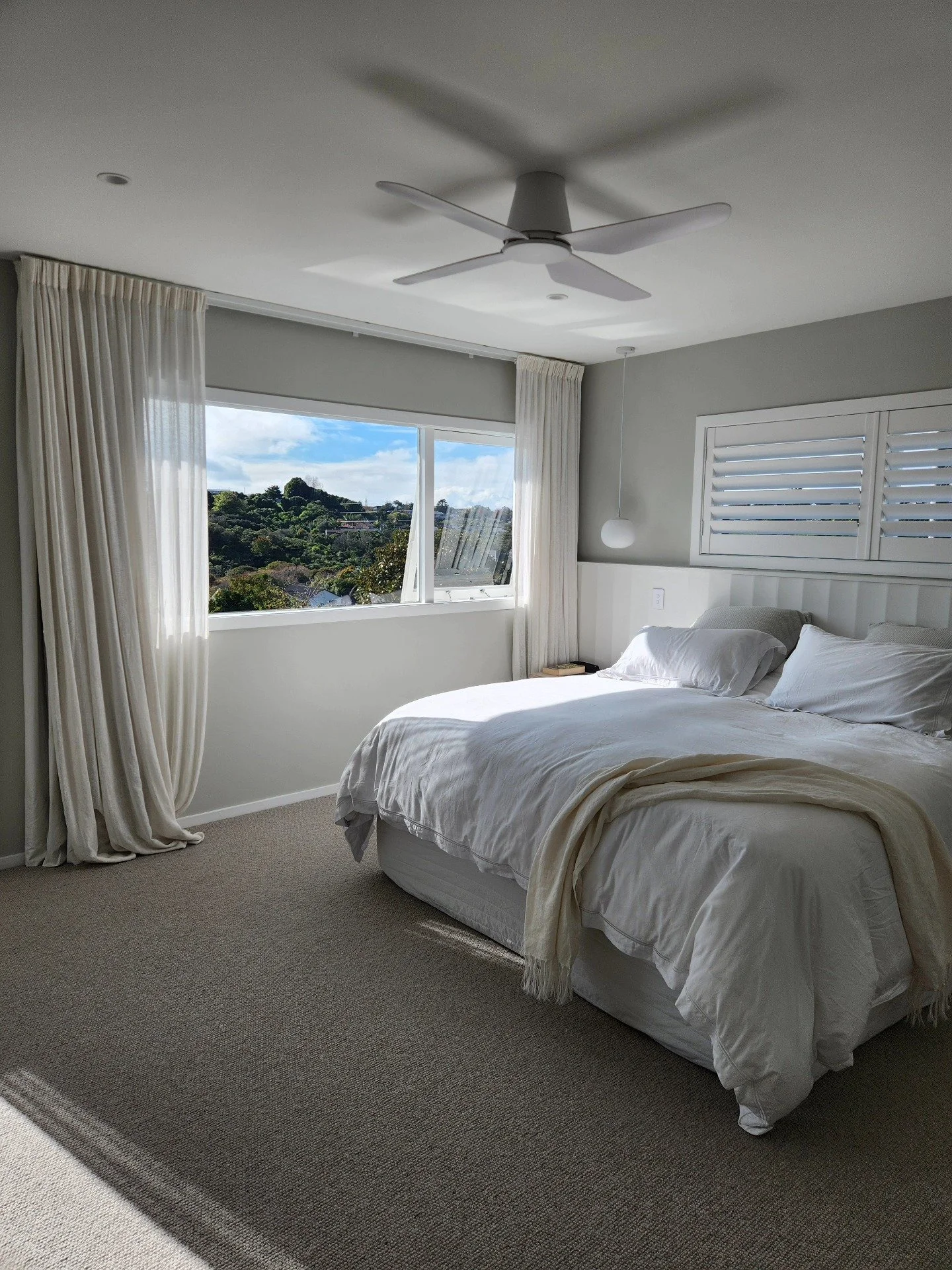 A bedroom with a large window showing a green hilly landscape. The bed is made with white linens, and there are white curtains on either side of the window. A small nightstand with a book and a lamp is next to the bed. The ceiling has a modern white fan, and there is a pendant light hanging over the bed.