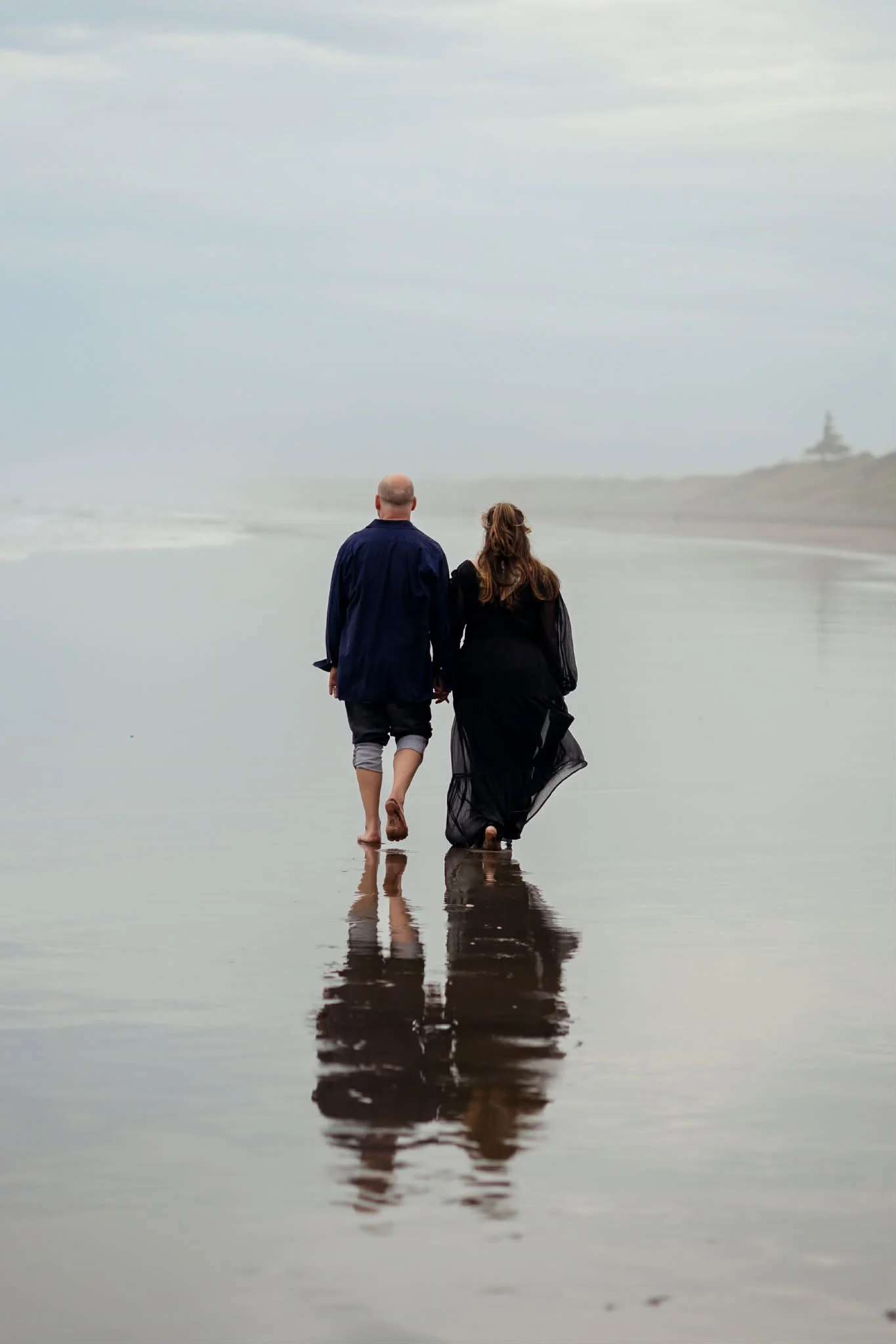 A man and woman walking barefoot on the beach at the water's edge during overcast weather, with their reflections visible on the wet sand, and a lighthouse in the distance.