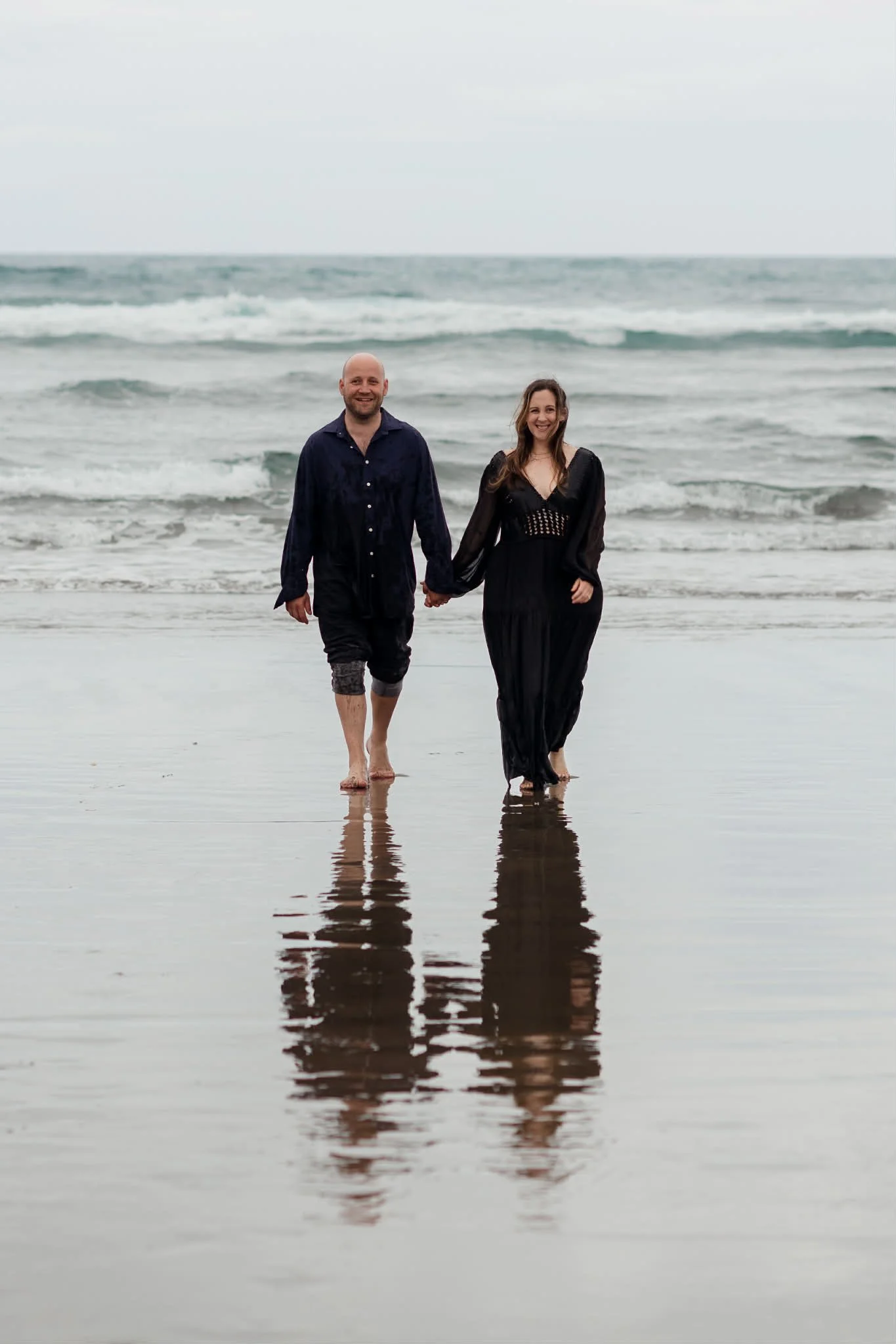 A smiling couple walking hand in hand on the beach with the ocean in the background and their reflection visible in the wet sand.