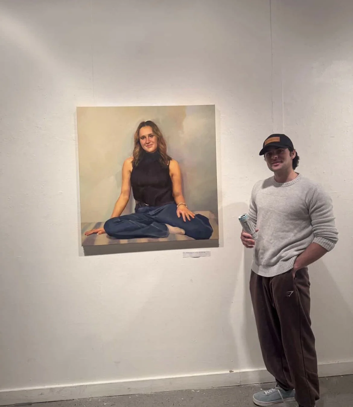 A young man in a gray sweater, brown pants, and a black cap standing beside a large portrait of a woman sitting on the floor in a black top and blue pants, with a neutral background, in an art gallery.