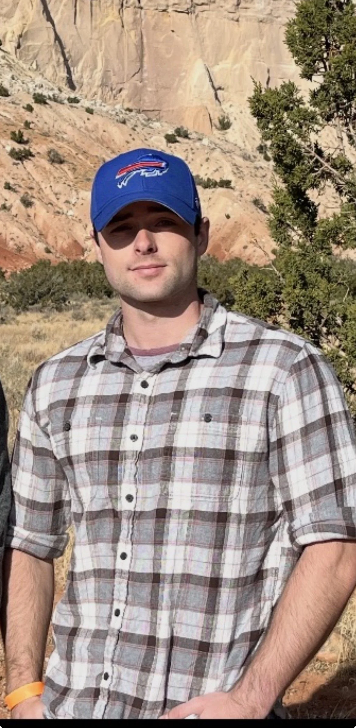 A man standing outdoors in front of rocky cliffs and some trees, wearing a blue baseball cap with a team logo and a plaid button-up shirt.