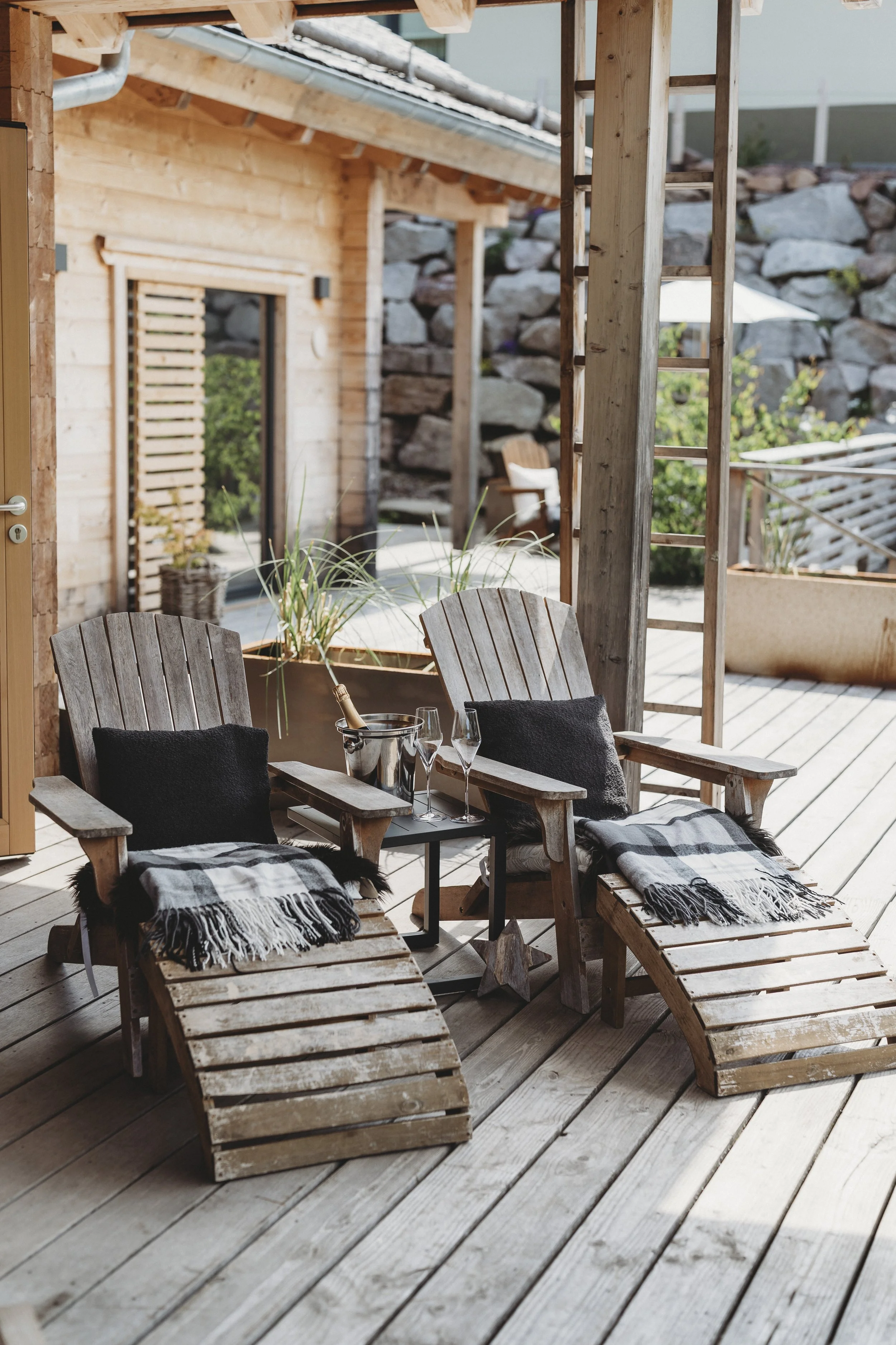 Ein Balkon mit zwei Holzliegen, dekoriert mit schwarzen Kissen und Plaids. Zwei Sektgläser, ein Flaschenkühler mit Wein und eine Flasche stehen auf einem kleinen Tisch. Im Hintergrund Holzelemente und ein Garten mit Steinen und Pflanzen.