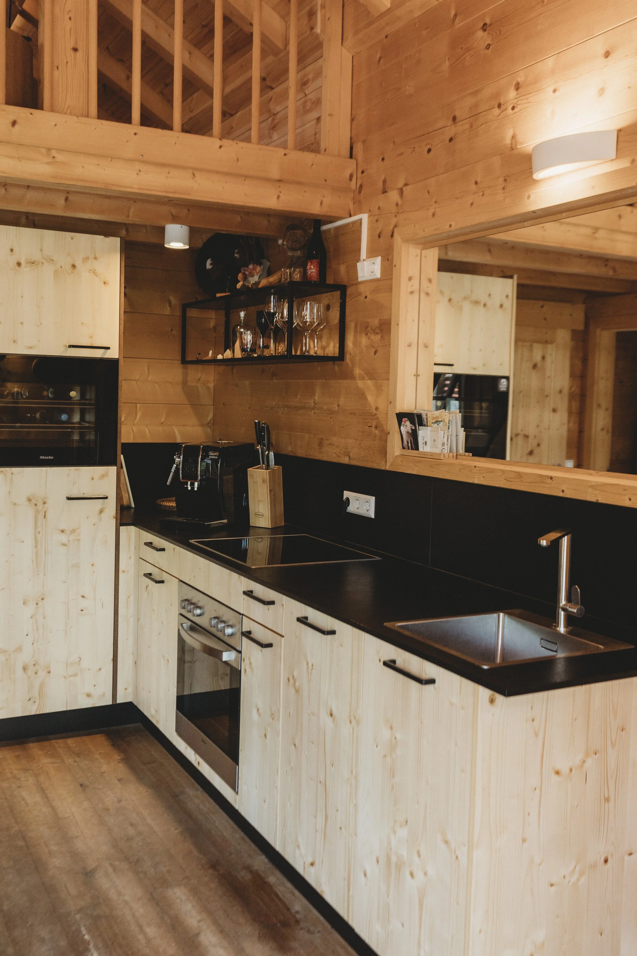 Der Blick auf eine Holz-Küche mit schwarzen Arbeitsflächen, einem Spülbecken, einem Herd und einem Spiegel, der den Raum reflektiert. Die Wände und die Decke bestehen aus Holzpaneelen, was eine gemütliche Atmosphäre schafft.