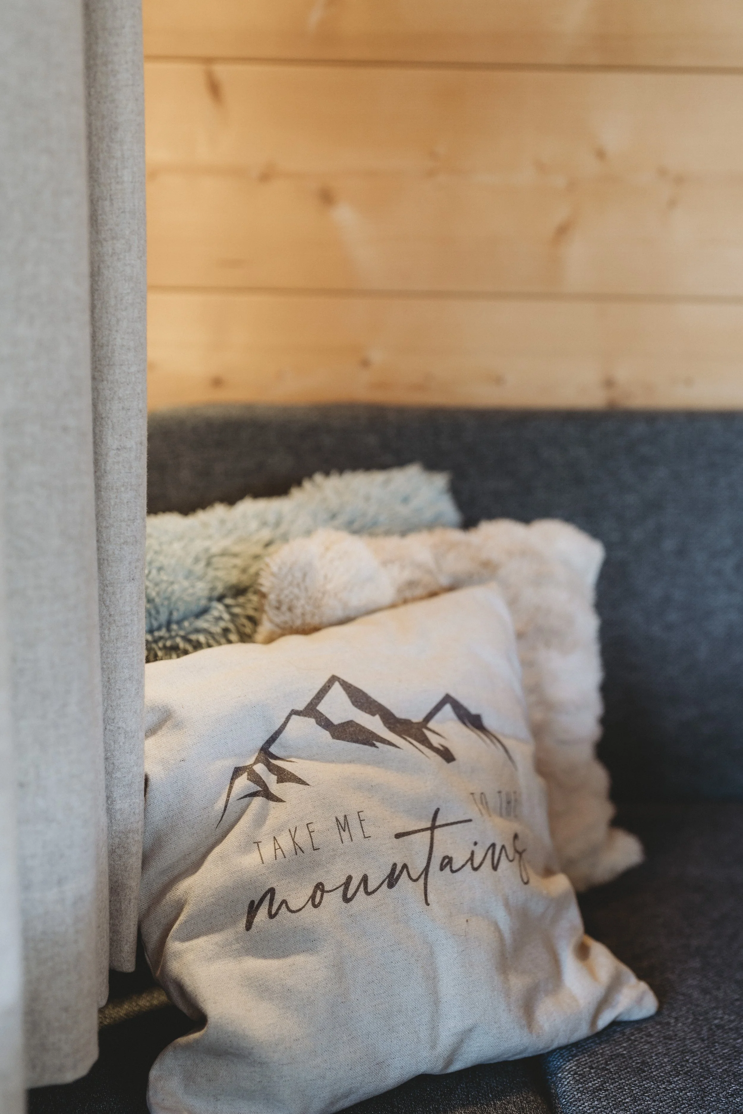 Ein Beutel aus Stoff mit Bergmotiven und dem Schriftzug "Take me to the mountains" liegt auf einem dunklen Oberflächen, im Hintergrund eine Holzpaneele und ein Bezug aus grauem Stoff.