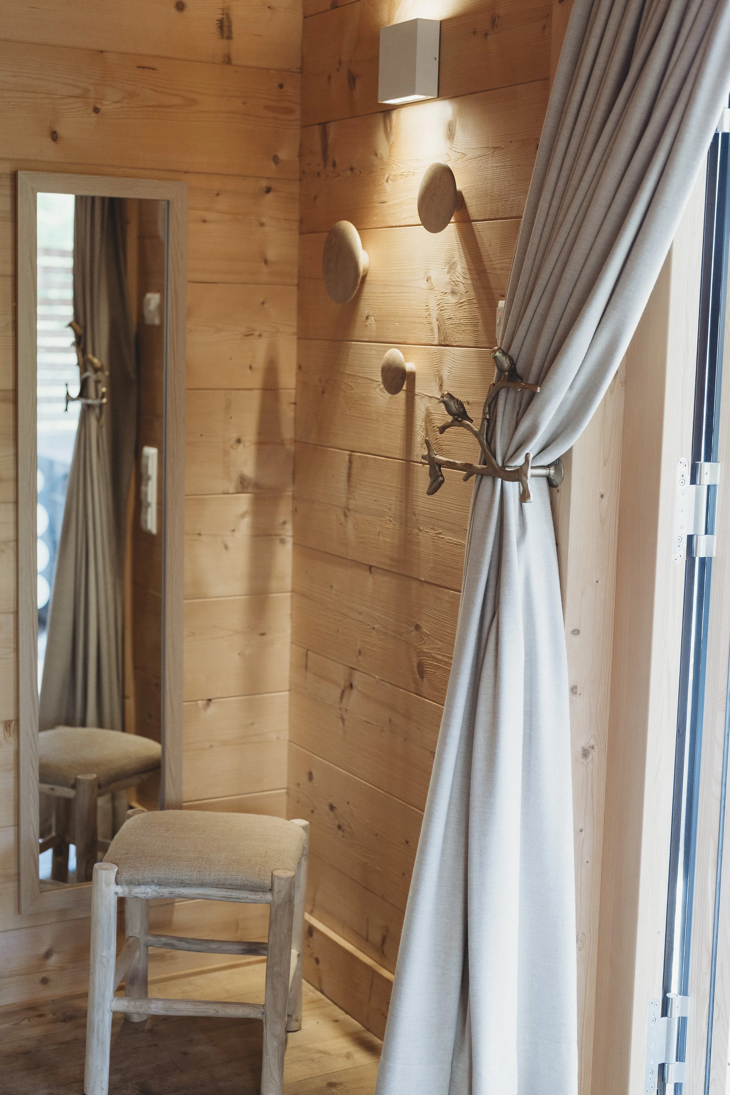 Ein Spiegel, ein Holzhintergrund mit Wanddekoration, ein Hocker mit beigeem Sitzbezug, ein Vorhang an einem Fenster, Wandlampen, die Licht auf die Holzplatte werfen.