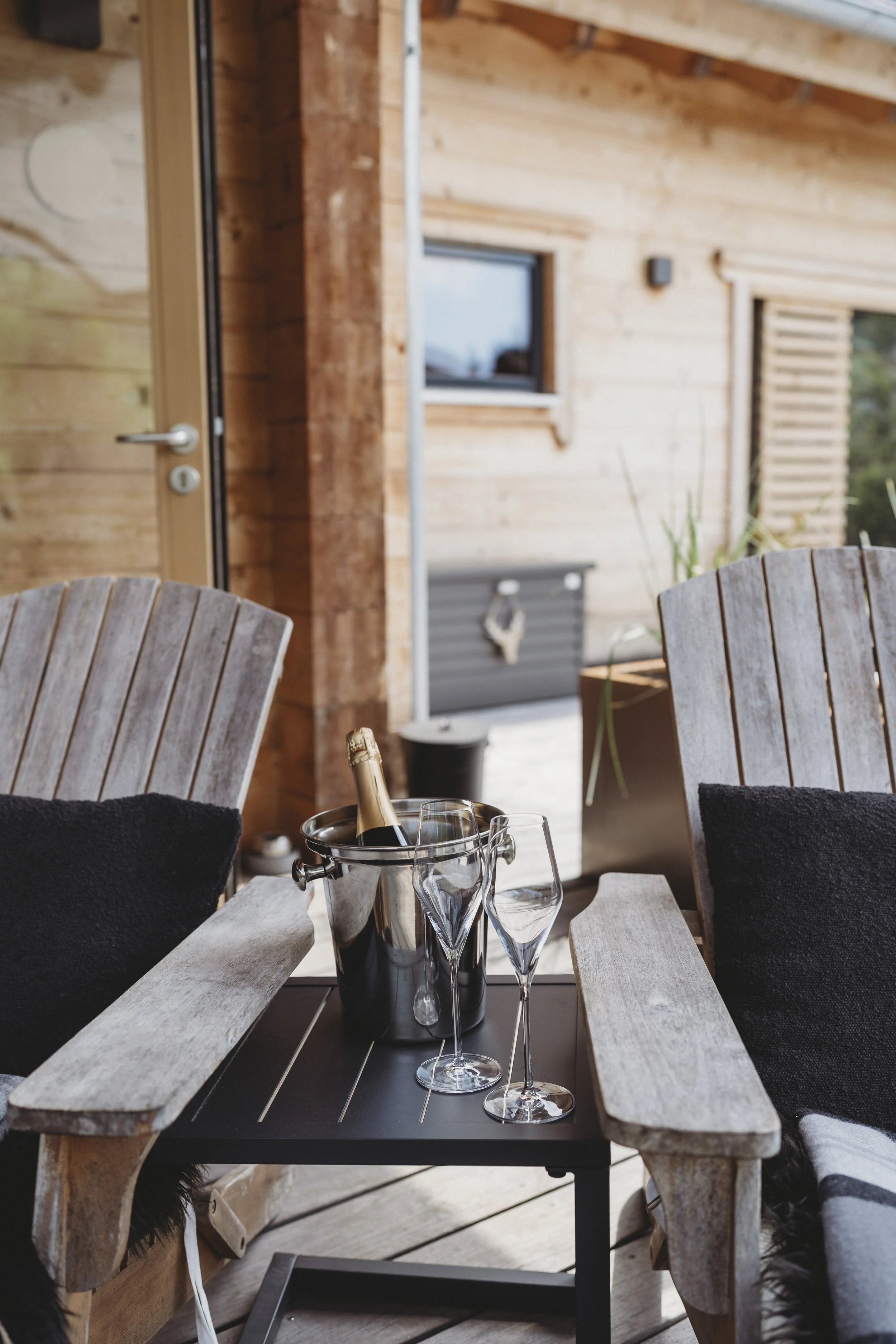 Ein Esstisch mit Champagnerflasche, Sektgläser, und Wein-Gläser im Vordergrund, umgeben von Holzmöbeln auf einer Terrasse mit Holzhaus-Hintergrund.