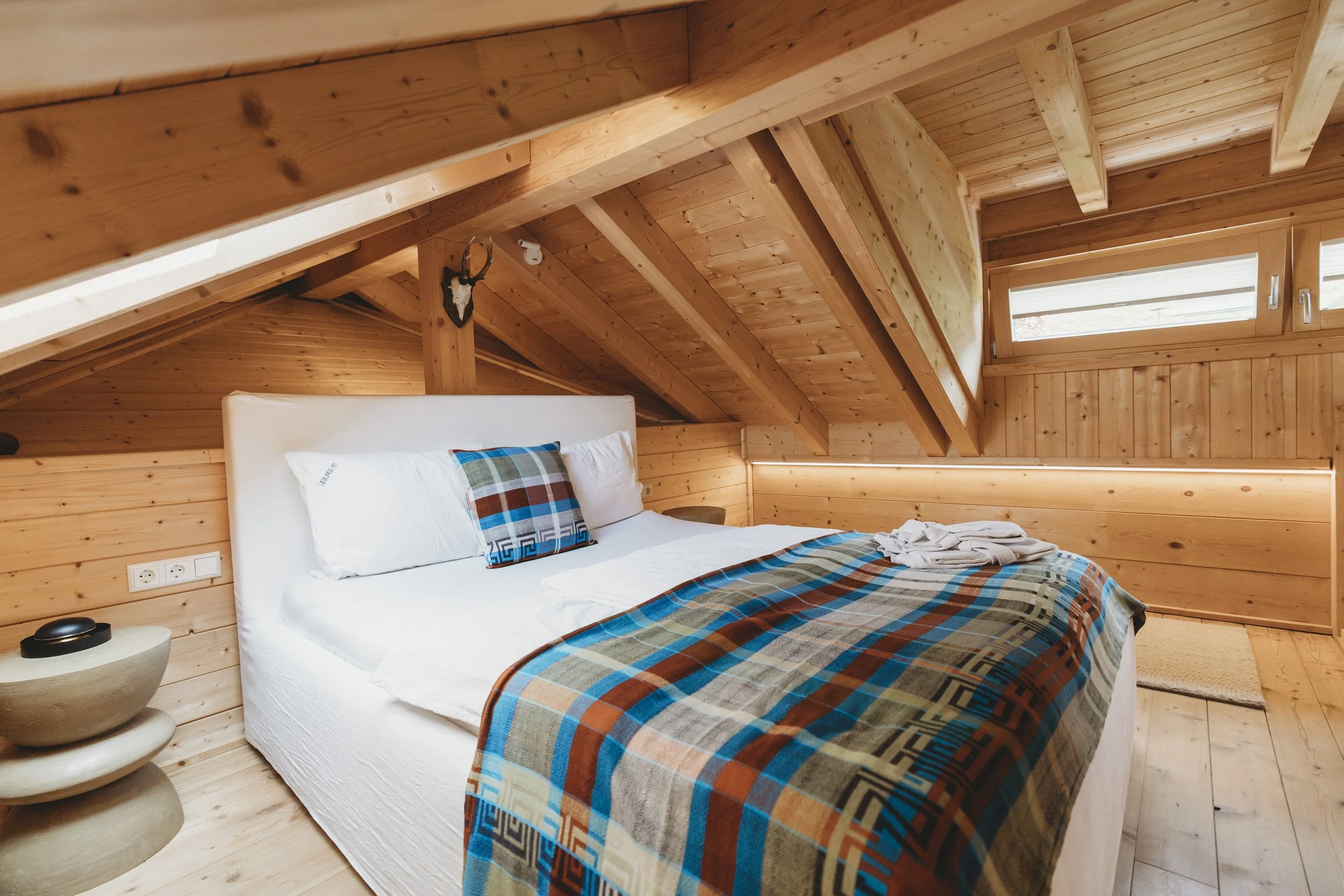 Ein Schlafzimmer mit einer Holzwand und -decke, einem weißen Bett mit buntem, kariertem Bettzeug, einem abgerundeten Nachttisch mit zwei Ablagen und einer Dekorationsharfe, sowie einem Fenster auf der rechten Seite.