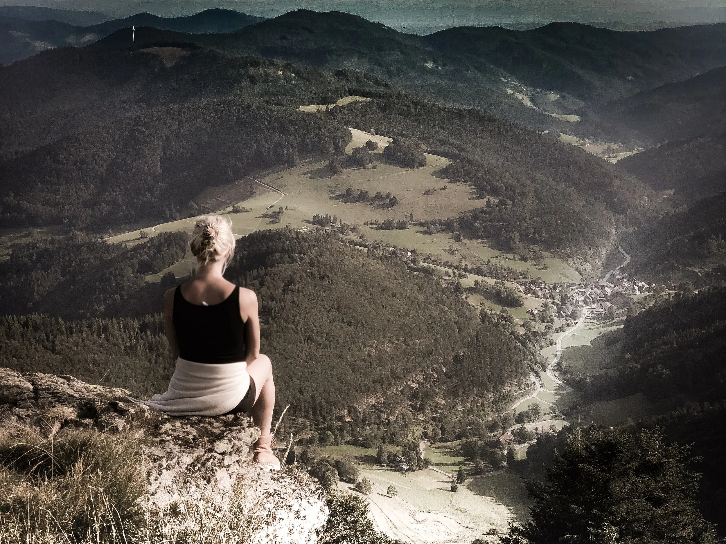 Eine Frau sitzt auf einem Felsen und blickt auf eine bergige Landschaft mit Wäldern, Wiesen und einer kleinen Stadt. Im Hintergrund sind Hügel und Berge zu sehen.