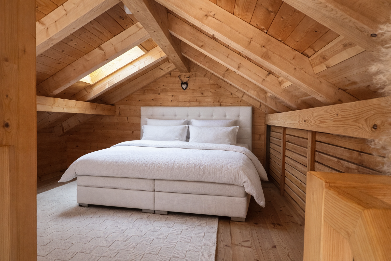 Schlafzimmer im Dachgeschoss mit Holzwänden und -decken, einem weißen Bett mit Kopfteil, mehreren weißen Kissen, einem beige Teppich und einem kleinen Fenster im Dach.