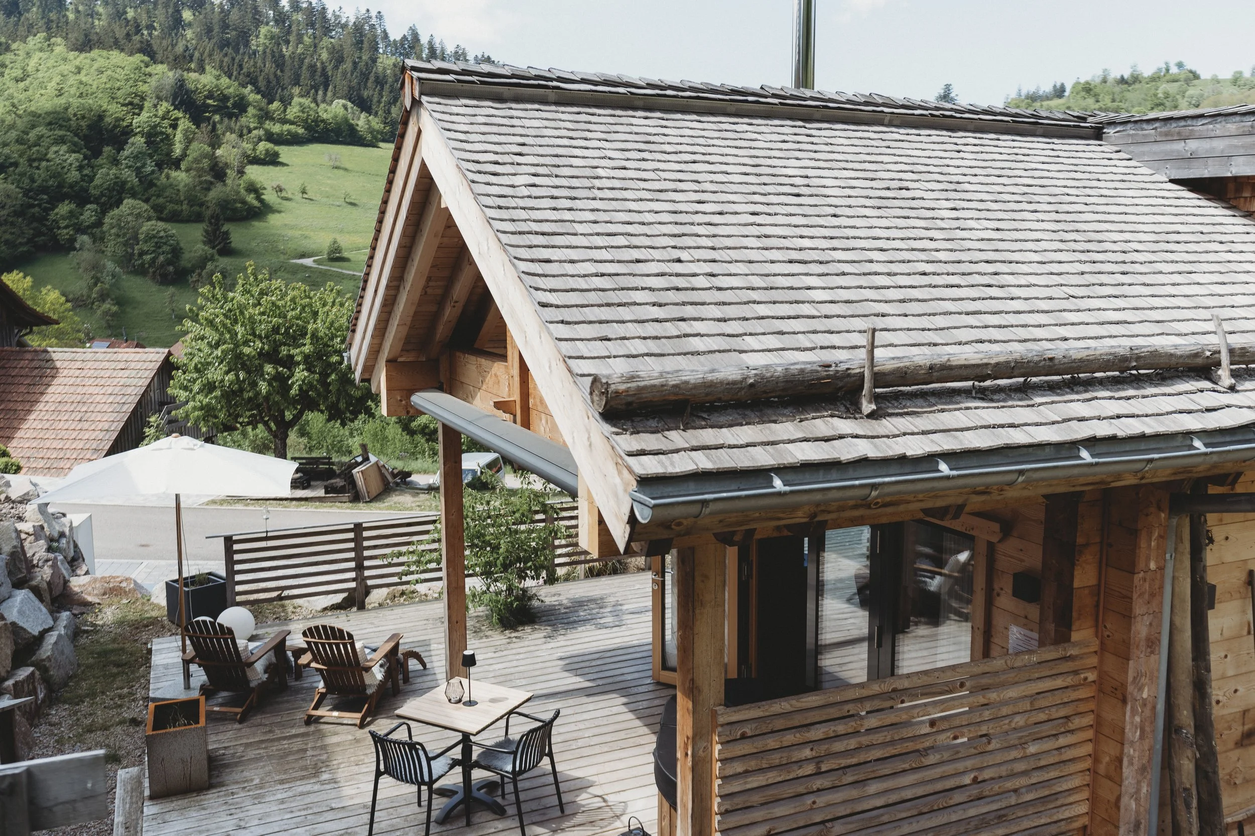 Das Bild zeigt ein Holzhaus mit einem Schindeldach, im Hintergrund grüne Hügel und Bäume. Im Vordergrund sind ein Terrassentisch mit Stühlen, ein Sonnenschirm und eine Terrasse sichtbar.