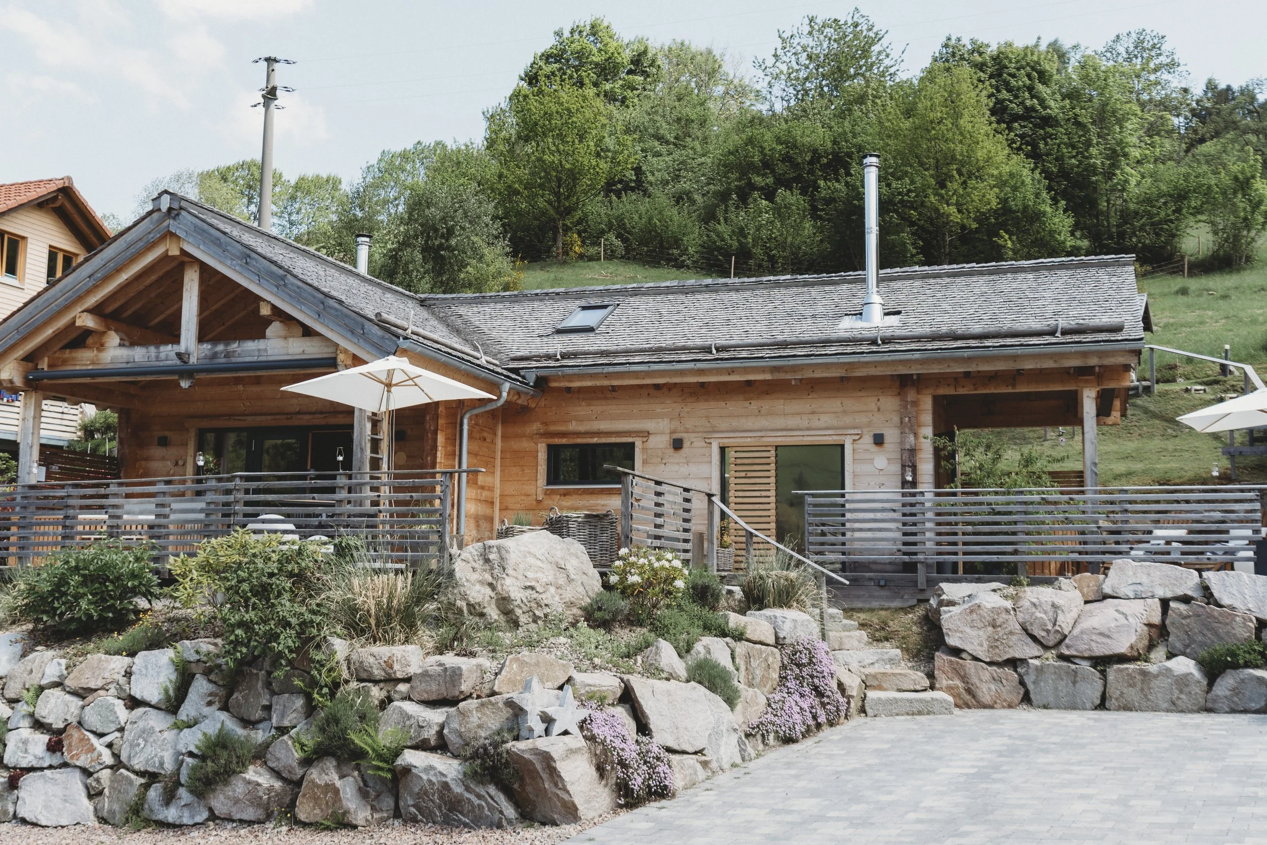 Ein Haus aus Holz mit Terrasse und Garten, umgeben von Steinen und Pflanzen, mit grünem Hügel im Hintergrund.