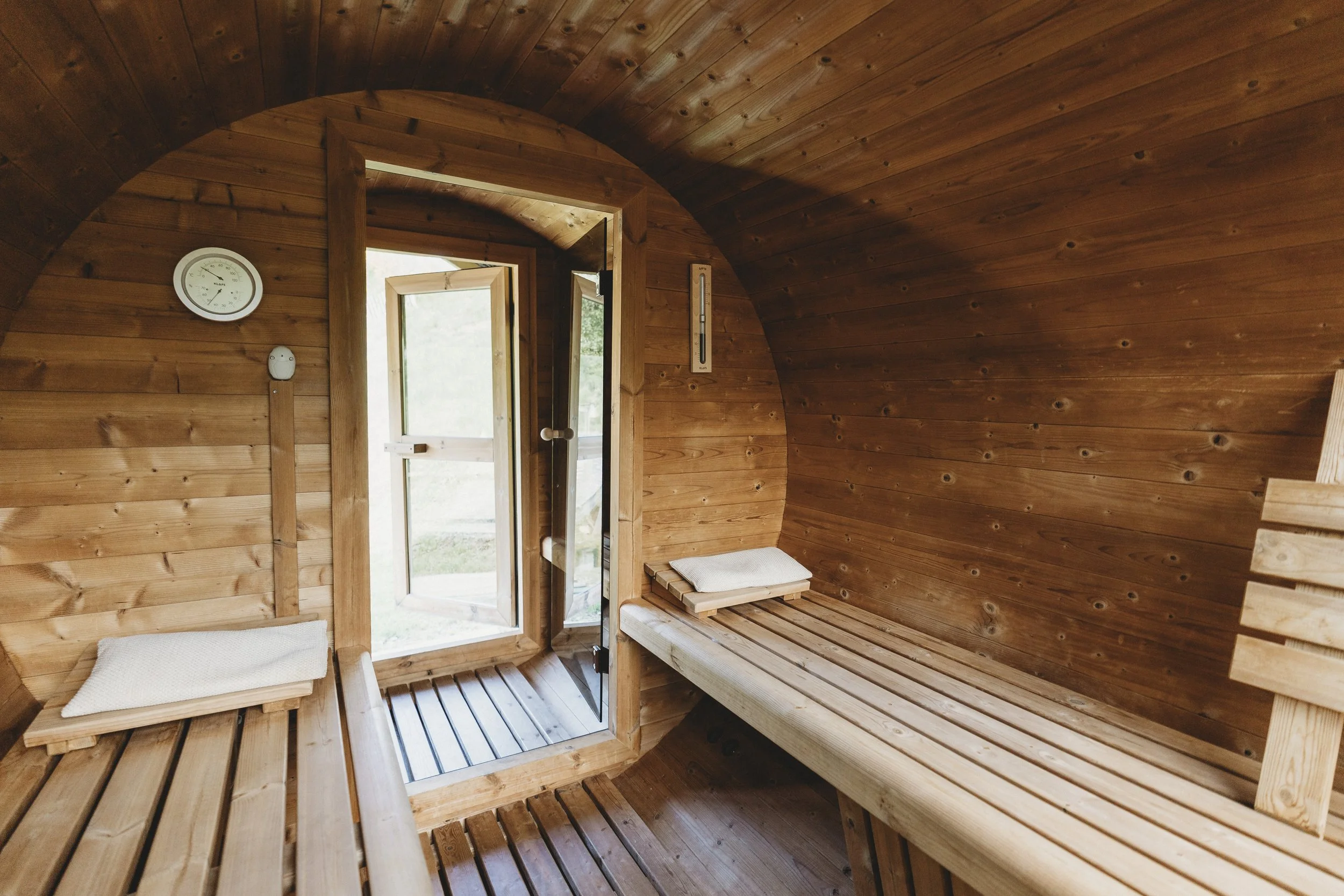 Innenraum einer Holzsauna mit Sitzbänken, Thermometer und Tür mit Glasfenster, natürlichem Licht und Holzvertäfelung