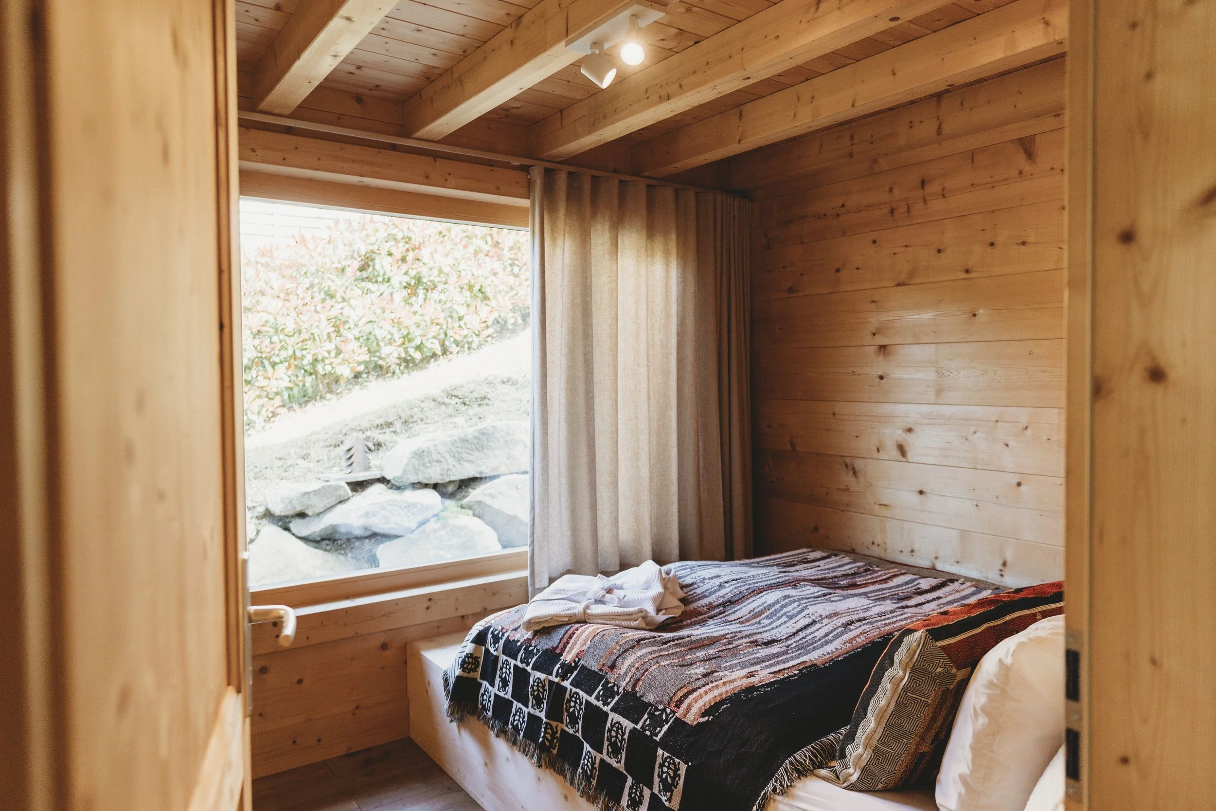 Ein Schlafzimmer mit Holzvertäfelung, ein Bett mit buntem Musterbettzeug, ein Fenster mit Blick ins Freie, Vorhänge und Kleidung auf dem Bett.