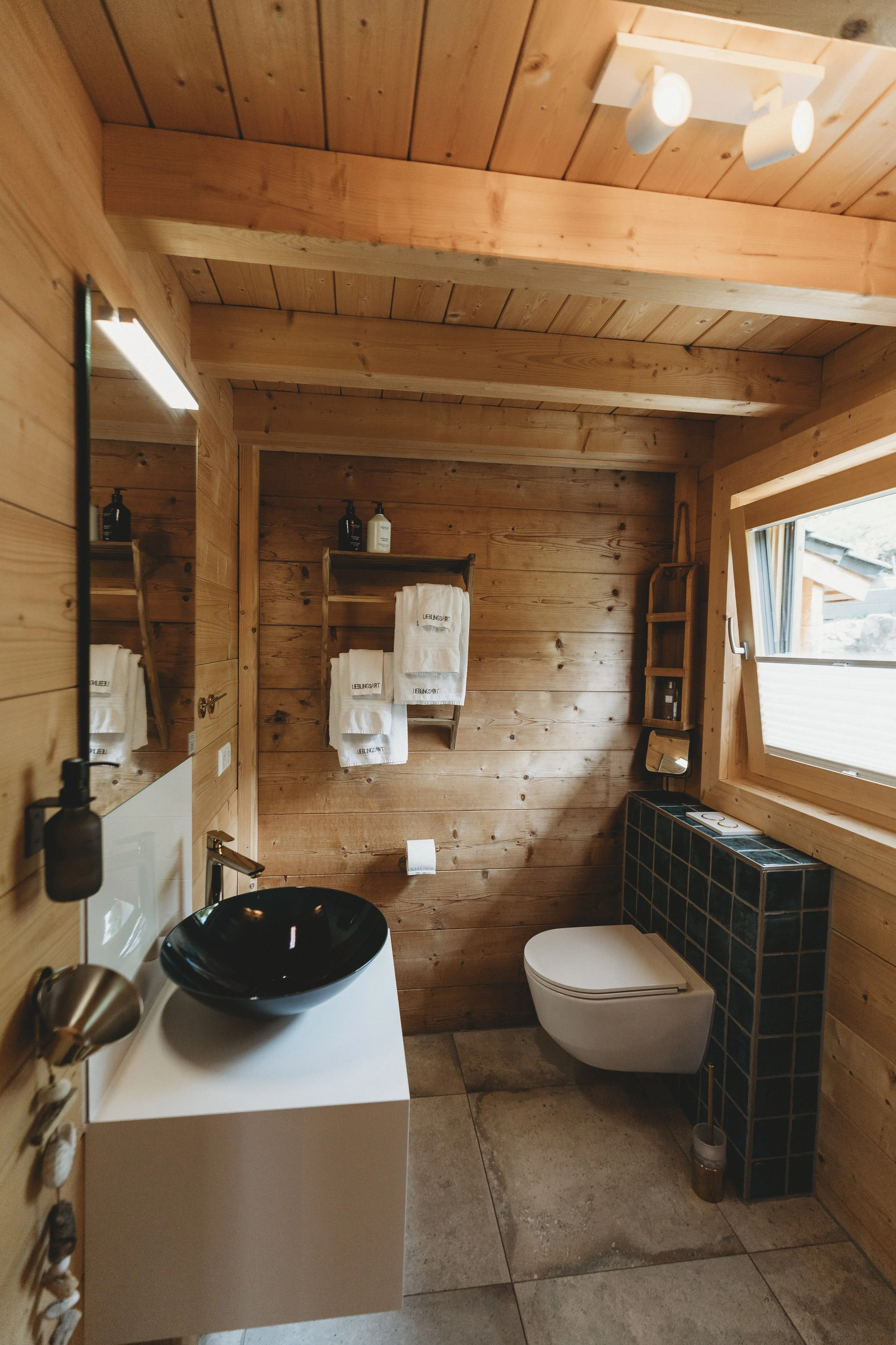 Das Bild zeigt ein modernes Badezimmer mit Holzverkleidung an Wänden und Decke. Es gibt einen schwarzen Waschtopf auf einer weißen Ablage, einen Spiegel, eine Toilette, eine schwarze Fliesensäule um die Toilette, einen Handtuchhalter mit weißen Handt