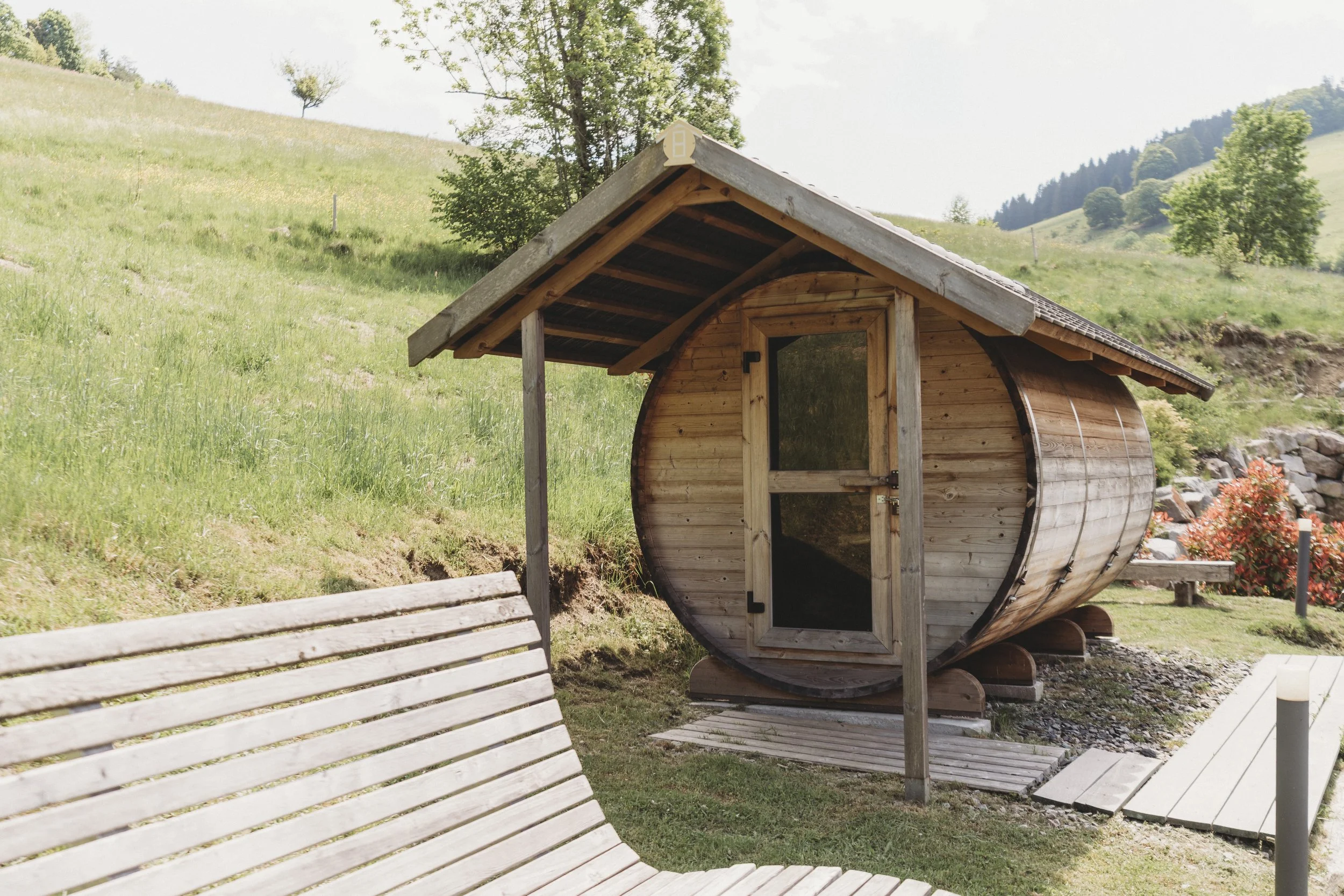 Ein kleines Holzhaus in Form einer aufgedrehten Trommel in einer grünen Berglandschaft mit Bäumen und Wiesen, im Vordergrund eine weiße Bank.
