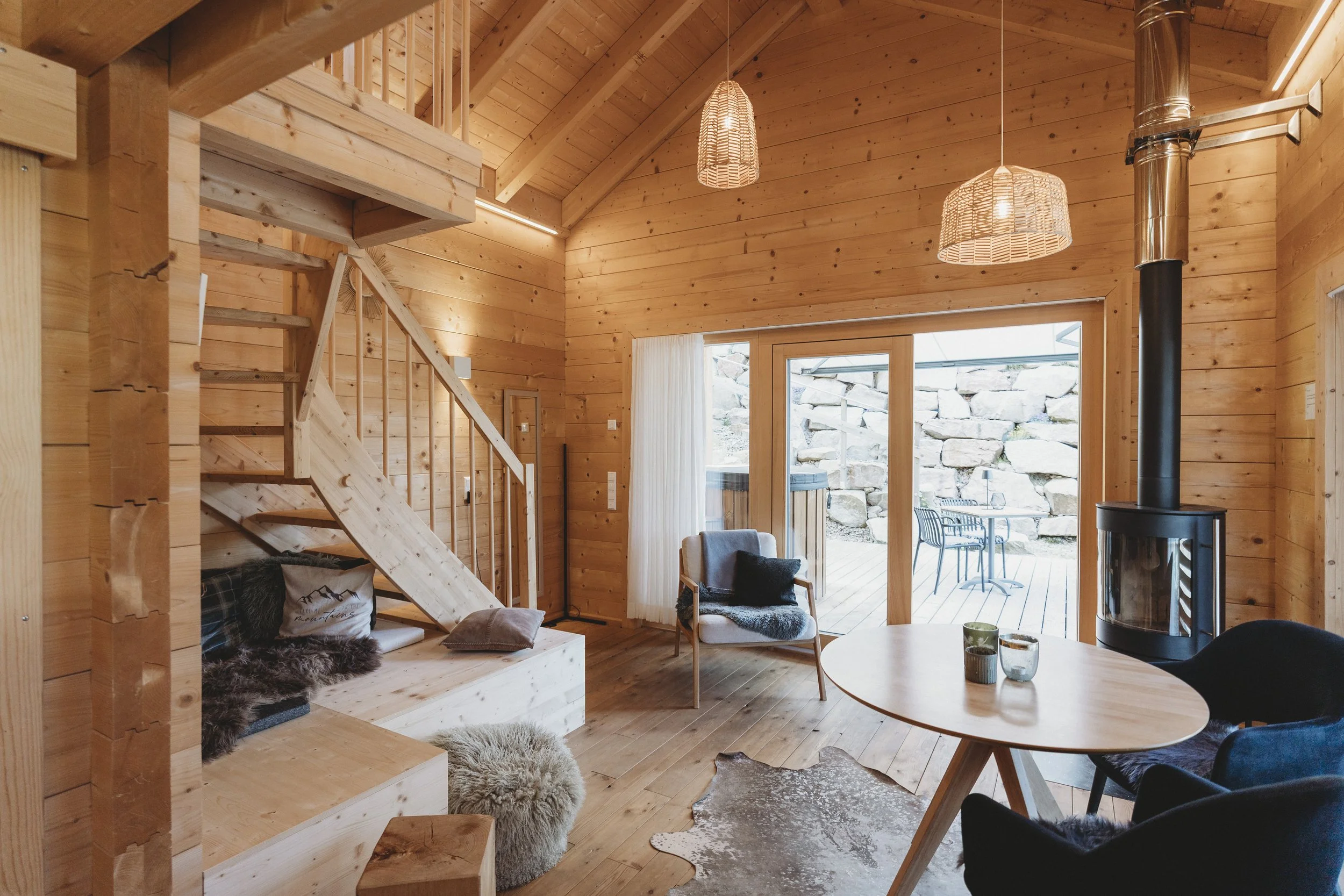 Innenansicht eines wohnlichen Holzhauses mit Lounge-Ecke, Kamin, großen Fenstern und Balkon im Hintergrund.