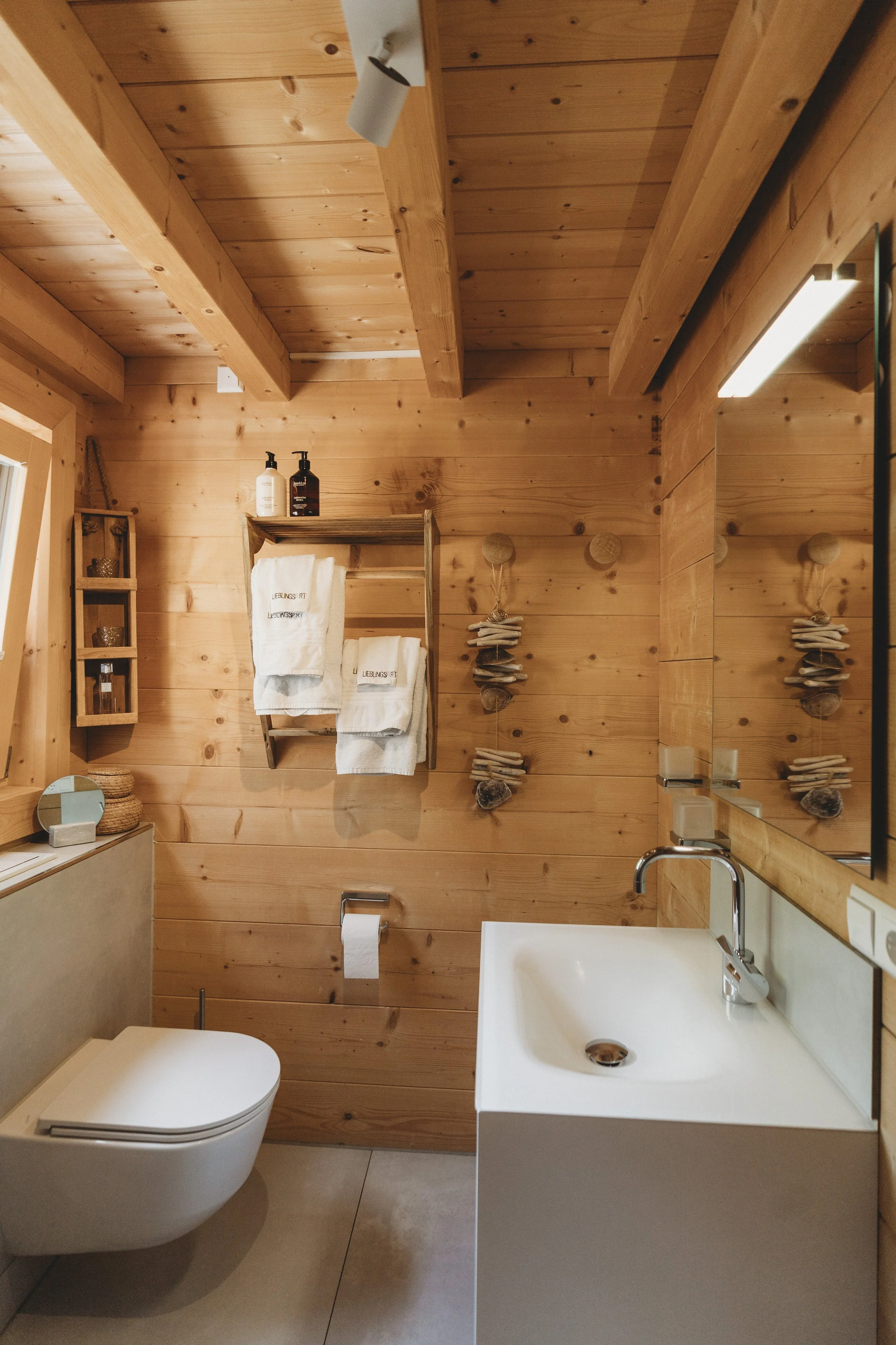 Ein Badezimmer mit Holzvertäfelung an Wänden und Decke, einem weißen Wand-WC und einem weißen Handwaschbecken. Es gibt Handtücher, Kerzen und Seifenspender. Spiegel und Beleuchtung sind an den Wänden angebracht.