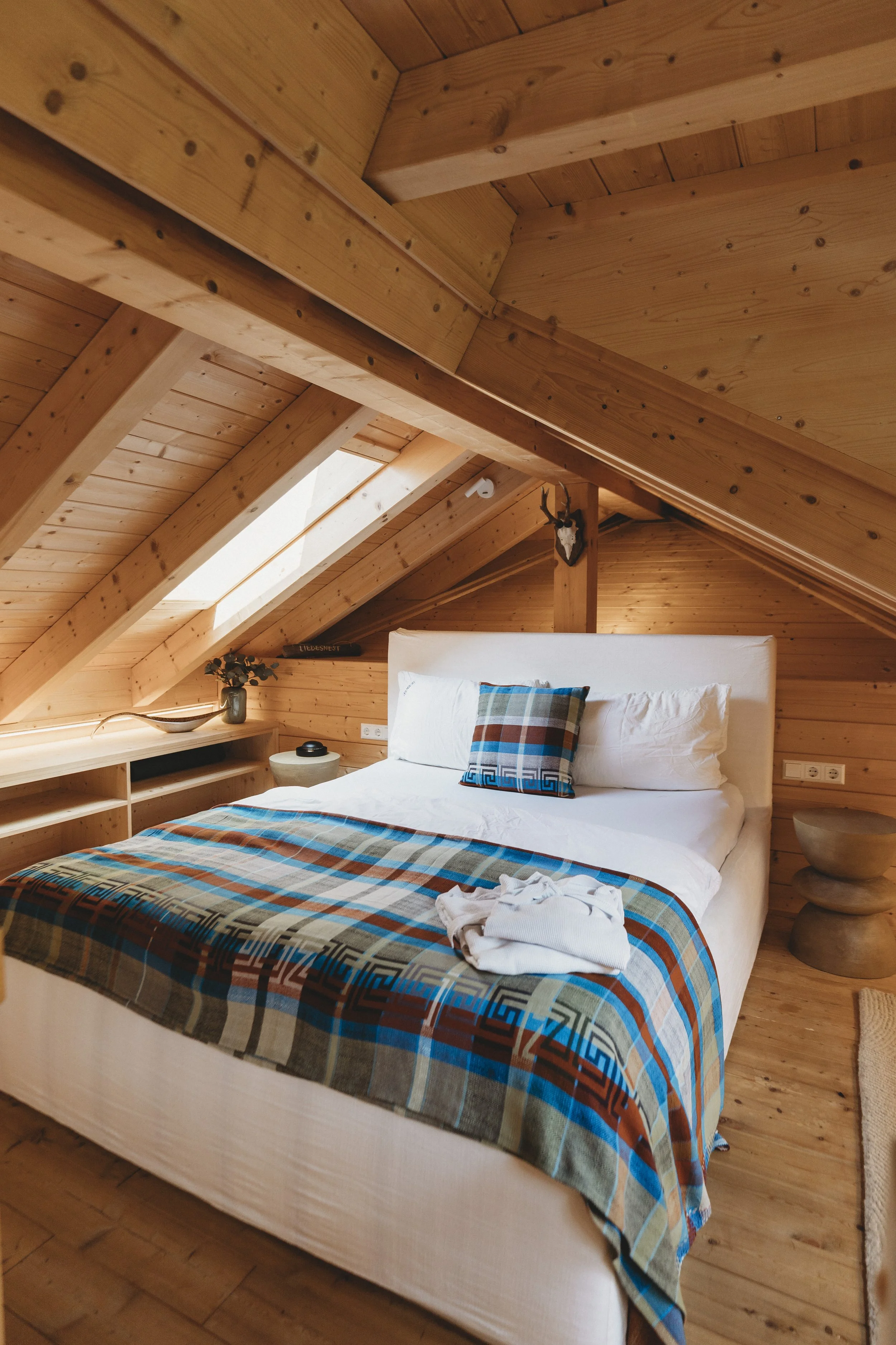 Ein Schlafzimmer mit einem weißen Bett, das mit buntem, geometrischem Bettzeug bedeckt ist, in einem Raum mit Holzdecke und -wänden. Es gibt eine Nachttischlampe, eine Pflanze und eine Deko an der Wand.