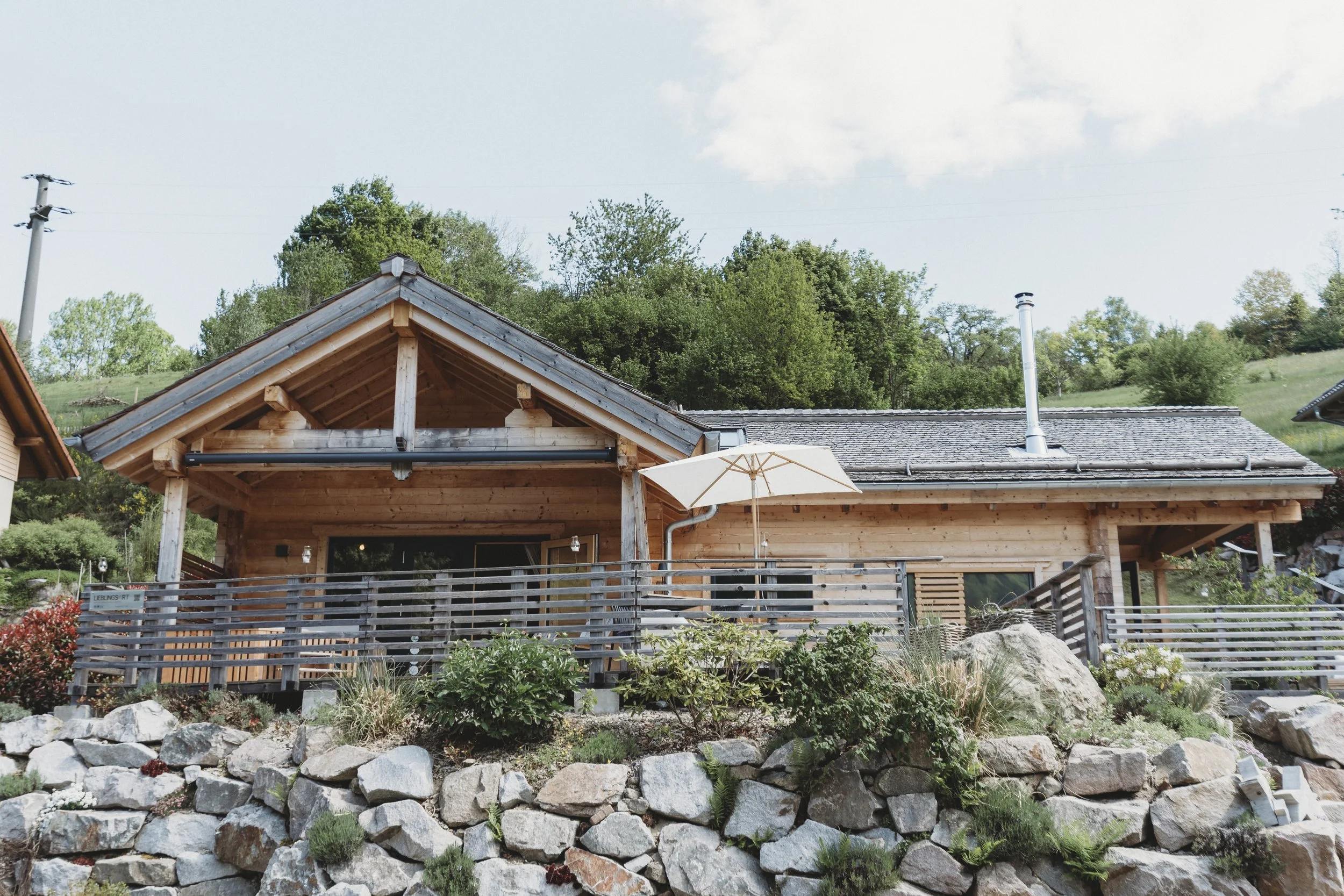 Ein Holzhaus mit Giebel, Schornstein und einer Gartenmauer, umgeben von Pflanzen und Bäumen, bei schönem Wetter.