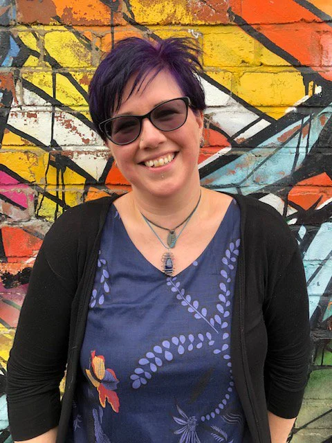A smiling woman with short purple hair and sunglasses stands in front of colorful graffiti on a brick wall.