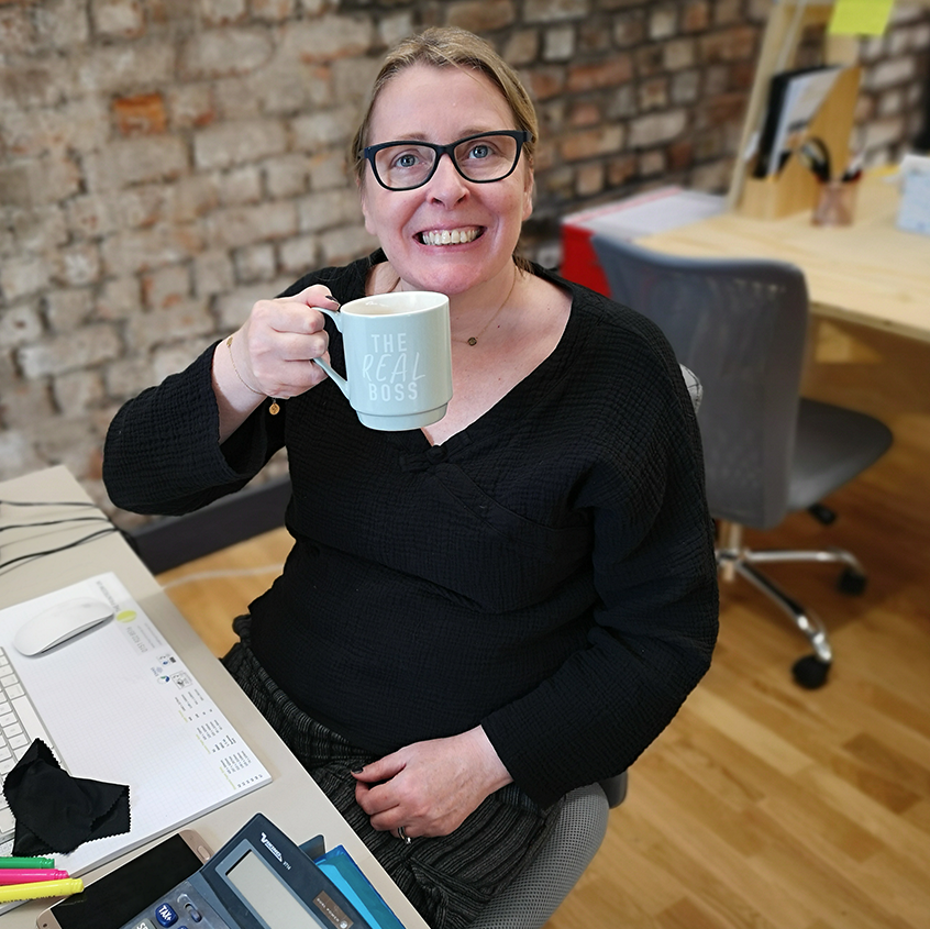 Image of a smiling woman with blonde hair and glasses. She is holding a pale blue mug saying the real boss.