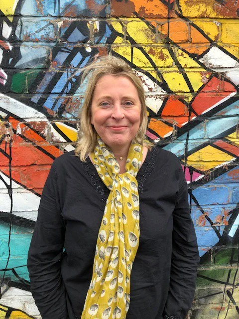 A woman with blonde hair wearing a black jacket and yellow scarf standing in front of a colorful graffiti mural on a brick wall.