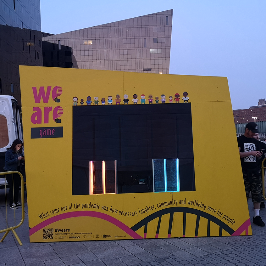 Image of the pod gamers could use to play the game on the side of the Museum of Liverpool. It's bright yellow with we are game written on it and the DNA strands in Pink and grey.