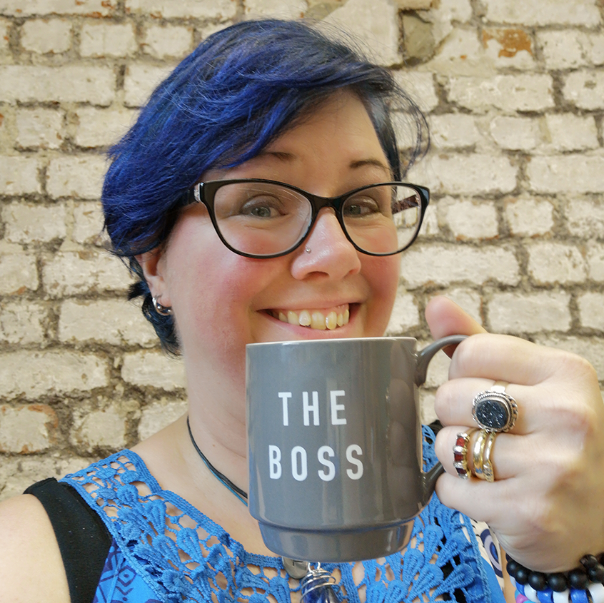 Image of a woman with short blue hair and glasses. She is smiling and holding a mug saying the boss.