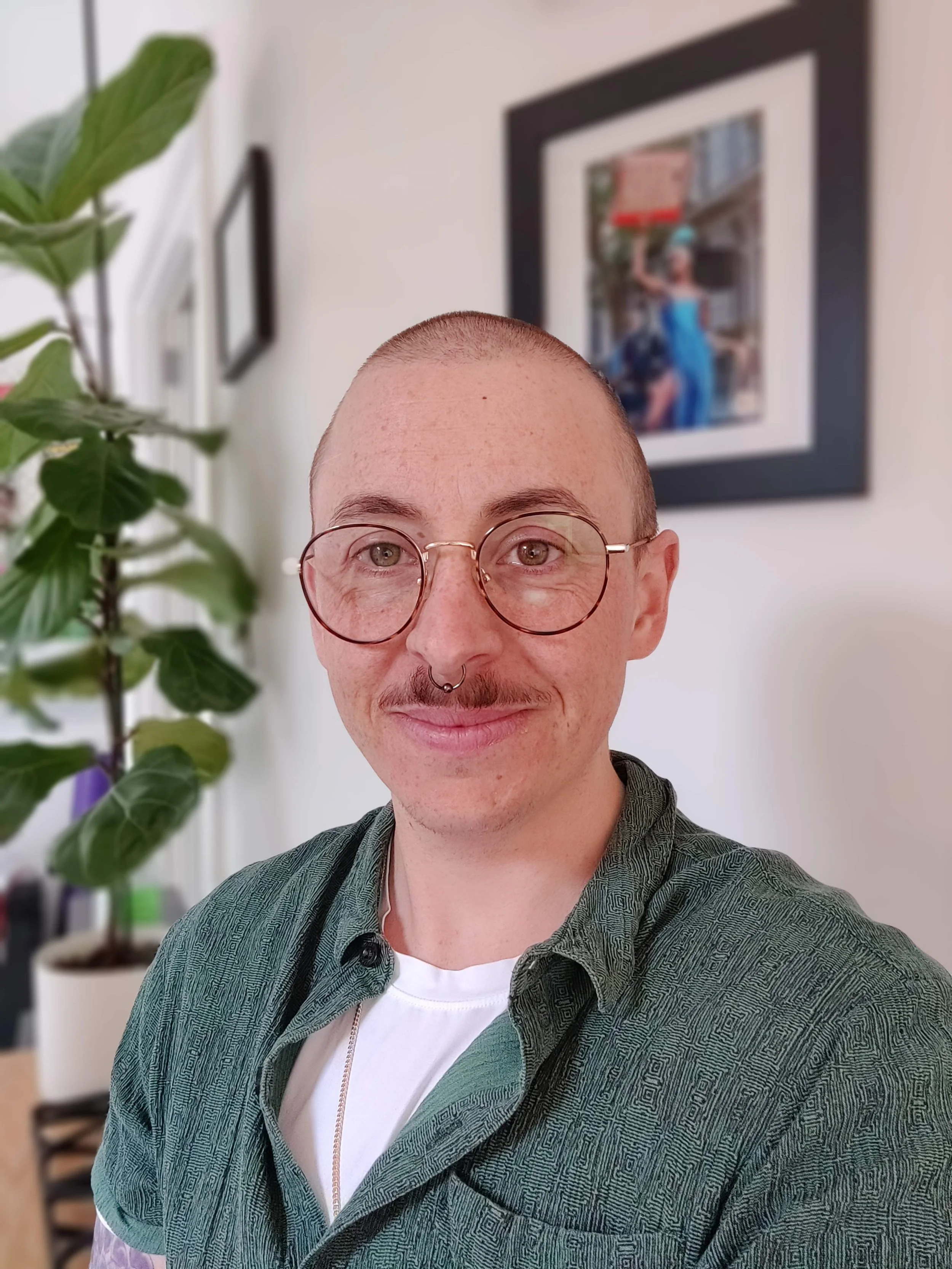 A person with a bald head, glasses, and a septum piercing smiling in a room with indoor plants and framed artwork on the wall.