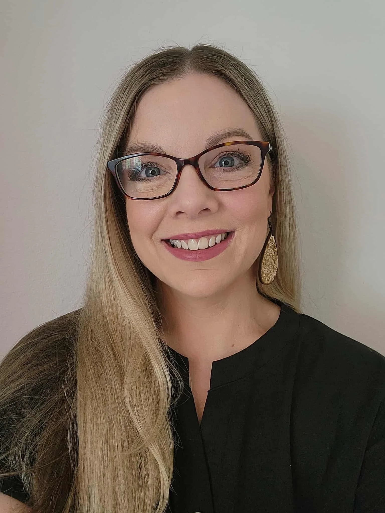 A woman smiling with long blond hair, wearing glasses, gold earrings, and a black top, standing against a plain light-colored wall.