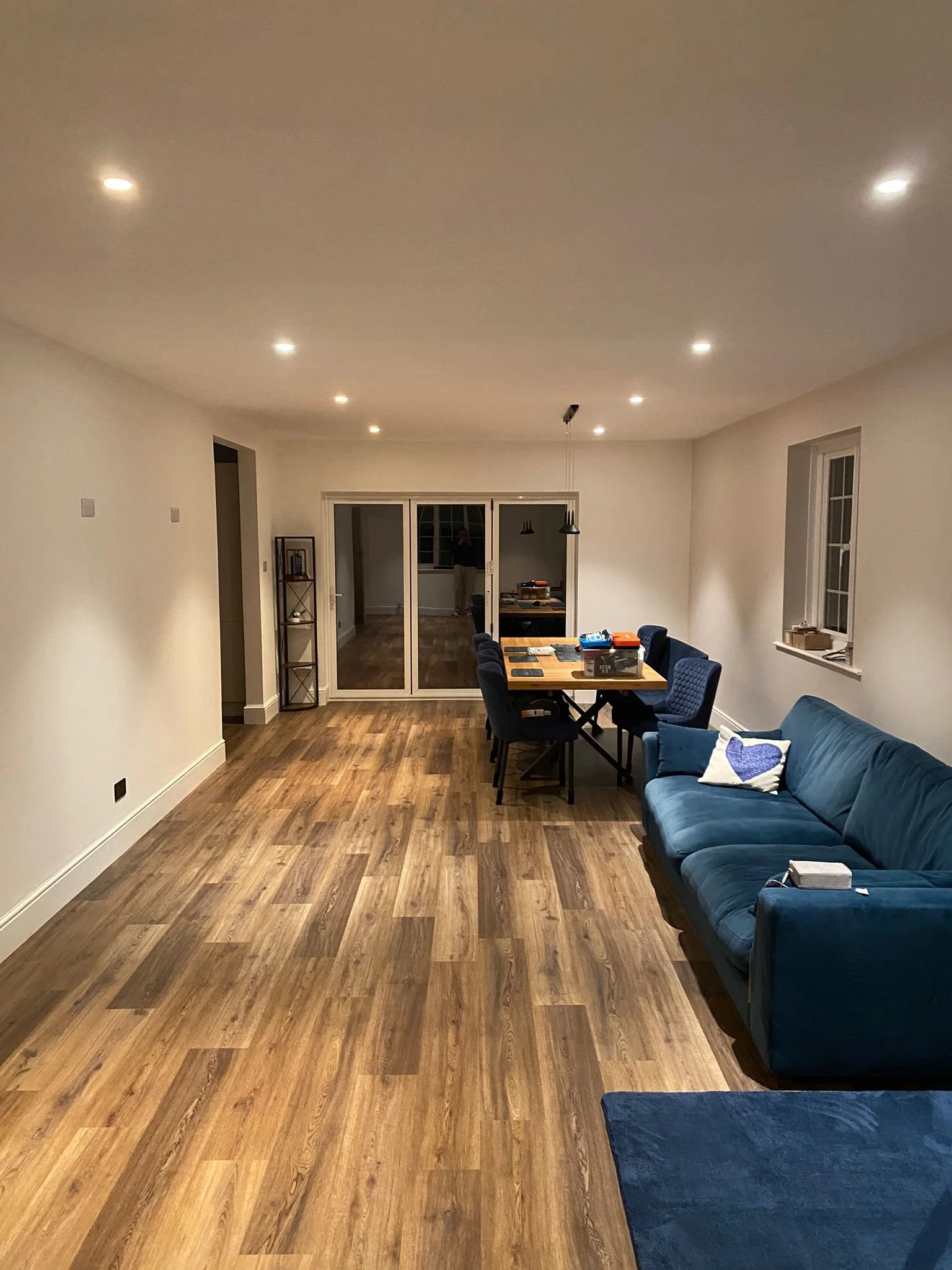 A spacious living room with wood flooring, when dusk outside, with a blue couch with decorative pillows, a dining table with chairs, a sliding glass door leading to a backyard, and overhead recessed lighting