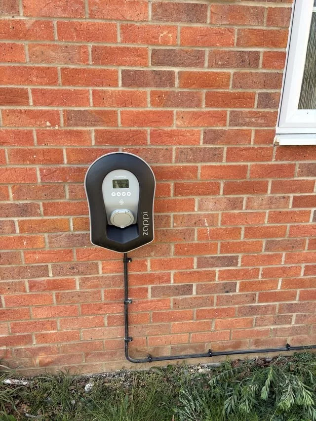 An electric vehicle charging station mounted on a brick wall outside a building with a black and gray design, and connected to underground wiring.