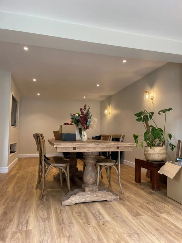 A dining room with a wooden table and six chairs, a large potted plant on a stool, and some boxes on the table. The room has wooden floors, white walls, and wall-mounted lights.