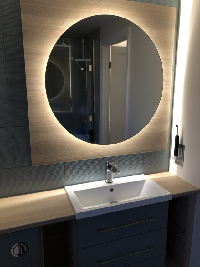 Modern bathroom vanity with a round mirror backlit with LED lighting, a white rectangular sink, and dark blue cabinets with horizontal handles. A toothbrush holder is mounted on the wall to the right of the mirror.