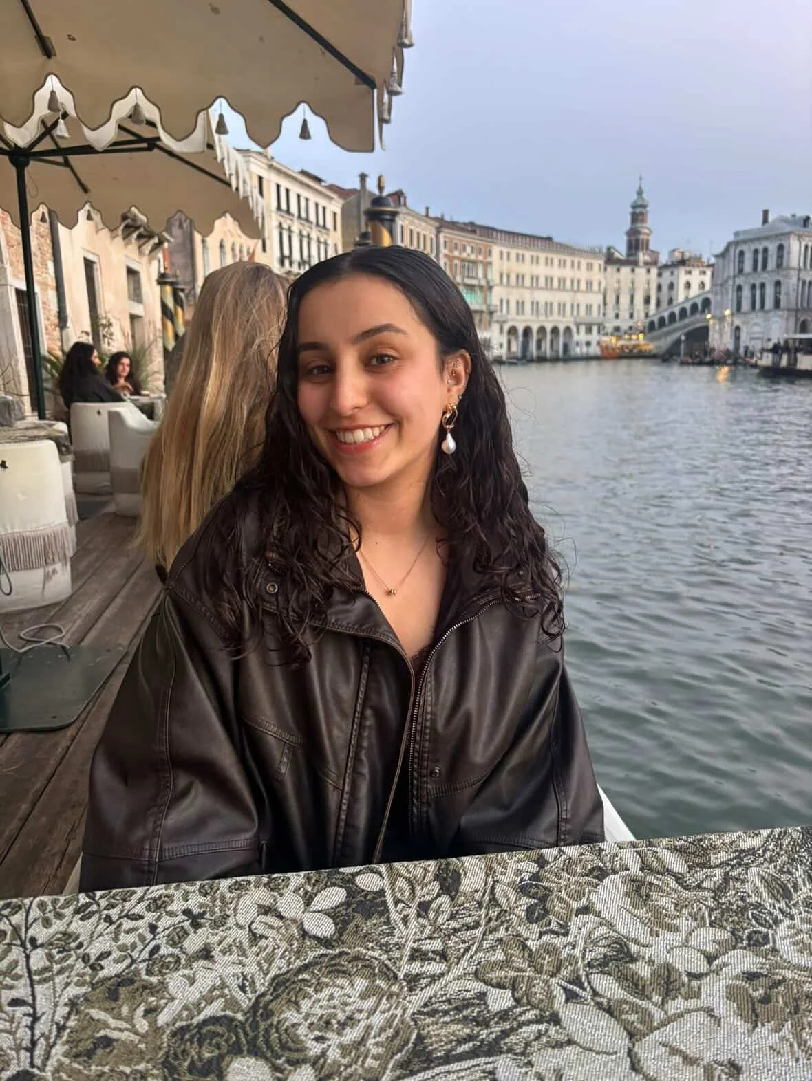 A young woman with dark, curly hair sitting at an outdoor restaurant table by a canal in Venice, Italy, with historic buildings and a bridge in the background during early evening.
