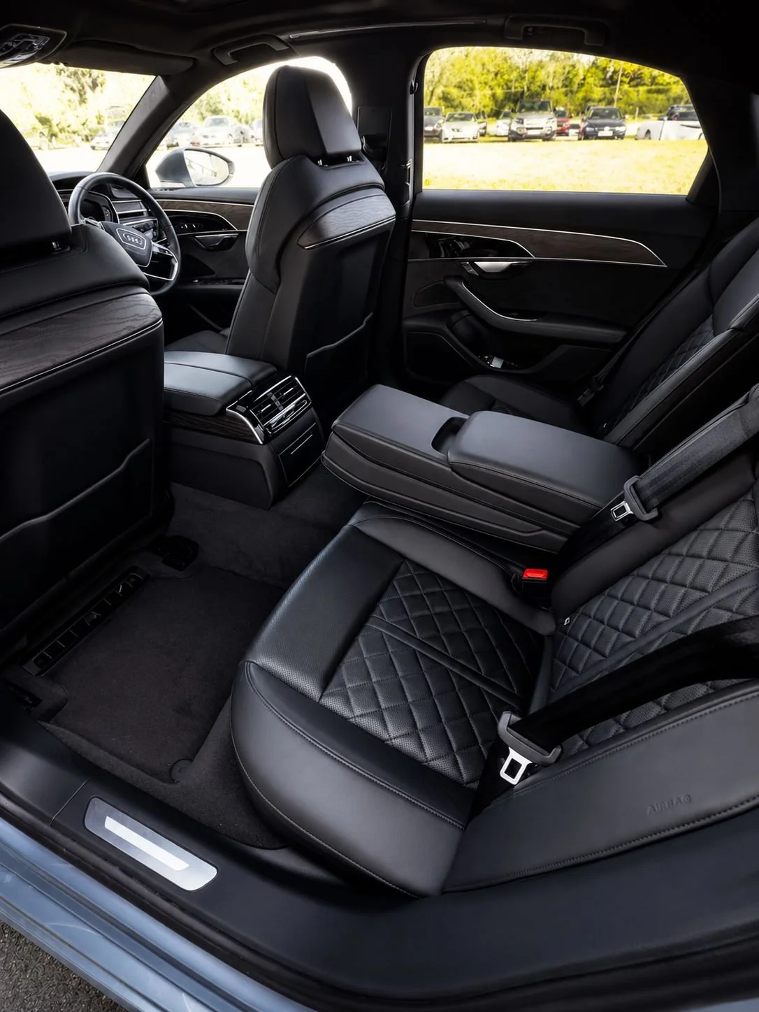Inside view of a luxury car's rear passenger area with black quilted leather seats, center armrest, and climate control vents, with a parking lot visible outside.