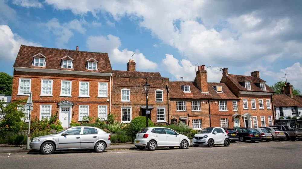 Row of traditional brick houses with pitched roofs, window dormers, cars parked along the street, and a cloudy sky.