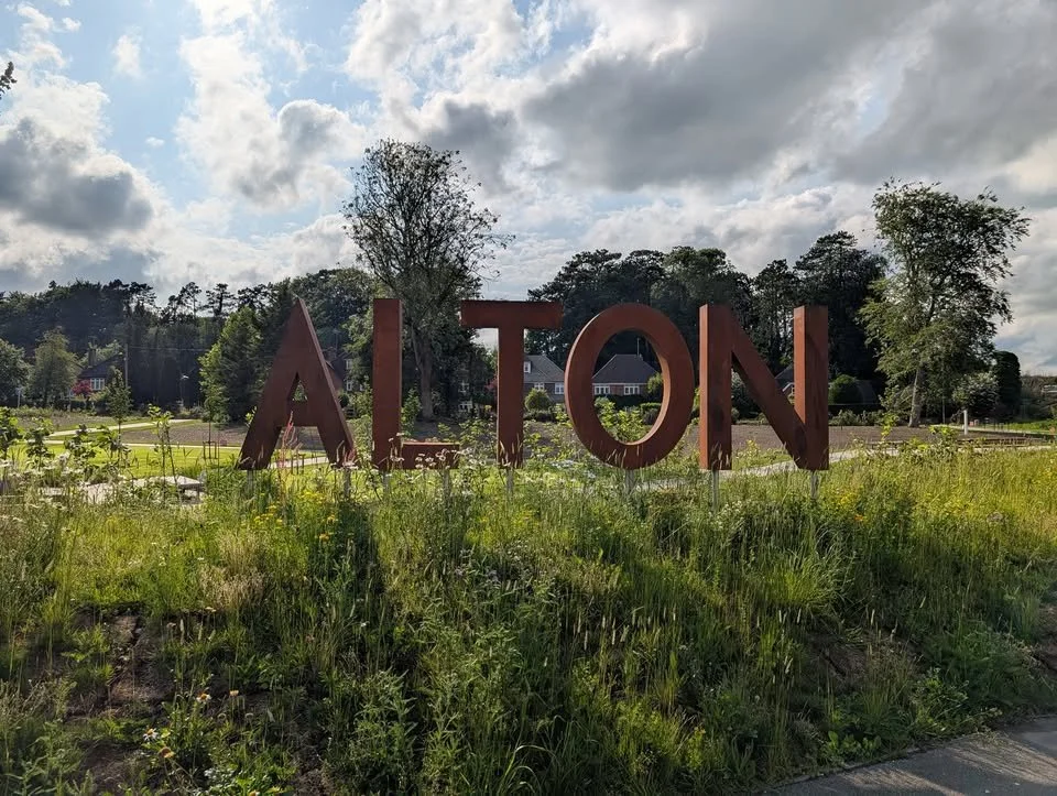 Large rust-colored metal letters spelling 'AITON' in a grassy area with trees and houses in the background under a partly cloudy sky.