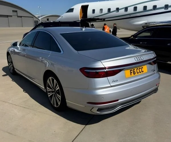 Silver Audi A8 luxury car parked on tarmac near private jet and hangar, with people in the background.