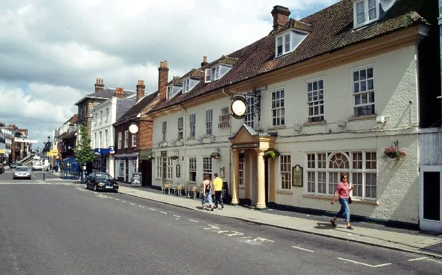 Street view of a row of historic buildings, including a hotel with columns and hanging flower baskets, with people walking on the sidewalk and cars parked along the street.