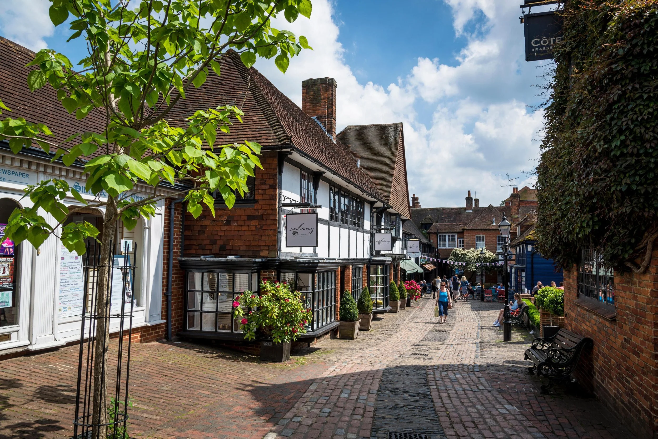 A bustling street scene in a small town with brick buildings, shops, and outdoor seating. People are walking and sitting, surrounded by greenery and flowering plants, under a partially cloudy sky.