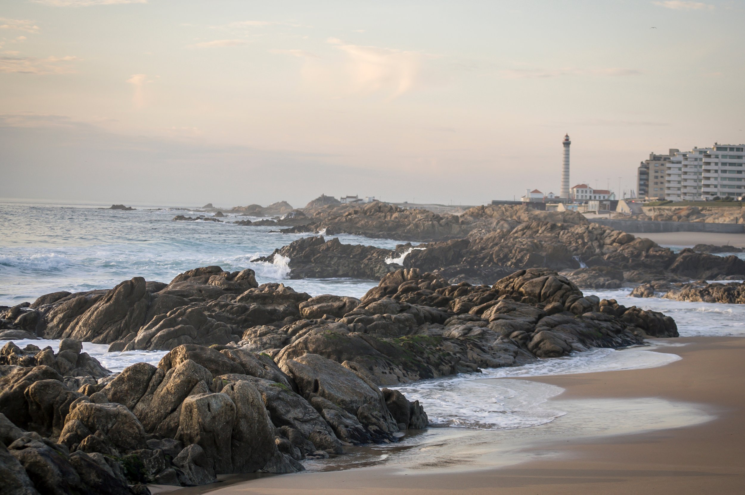 A rocky shoreline with waves crashing against the rocks, a sandy beach in the foreground, and a lighthouse with nearby buildings in the background under a partly cloudy sky.