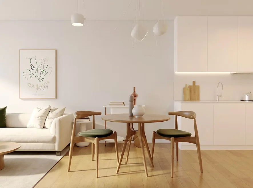 Modern, minimalist dining and living area with a round dining table, three chairs, white sofa with pillows, abstract artwork, and a white kitchen in the background.